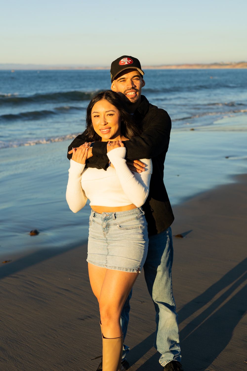 Couple embracing on a sunny beach, woman in white crop top and denim skirt, man in black jacket and cap.