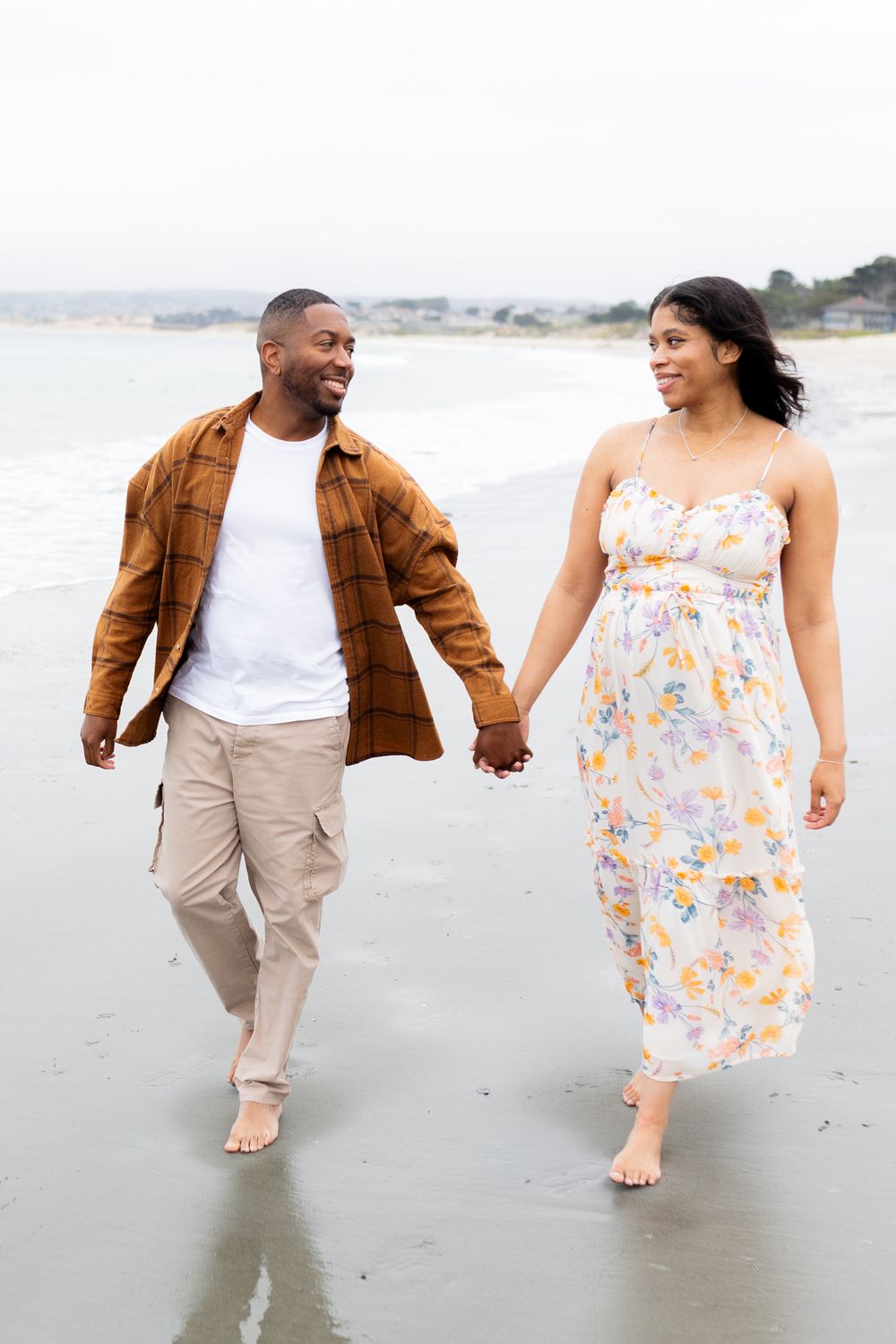 A happy couple walks hand-in-hand barefoot on a beach, the woman in a floral dress and the man in a brown shirt.