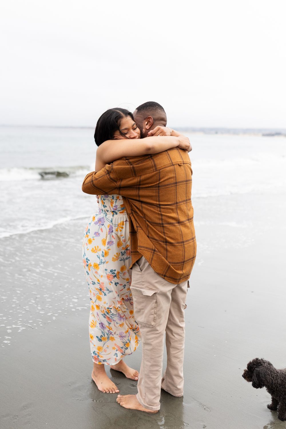 Couple embracing on a beach shore, woman in floral dress and man in plaid shirt, with a small dog nearby.