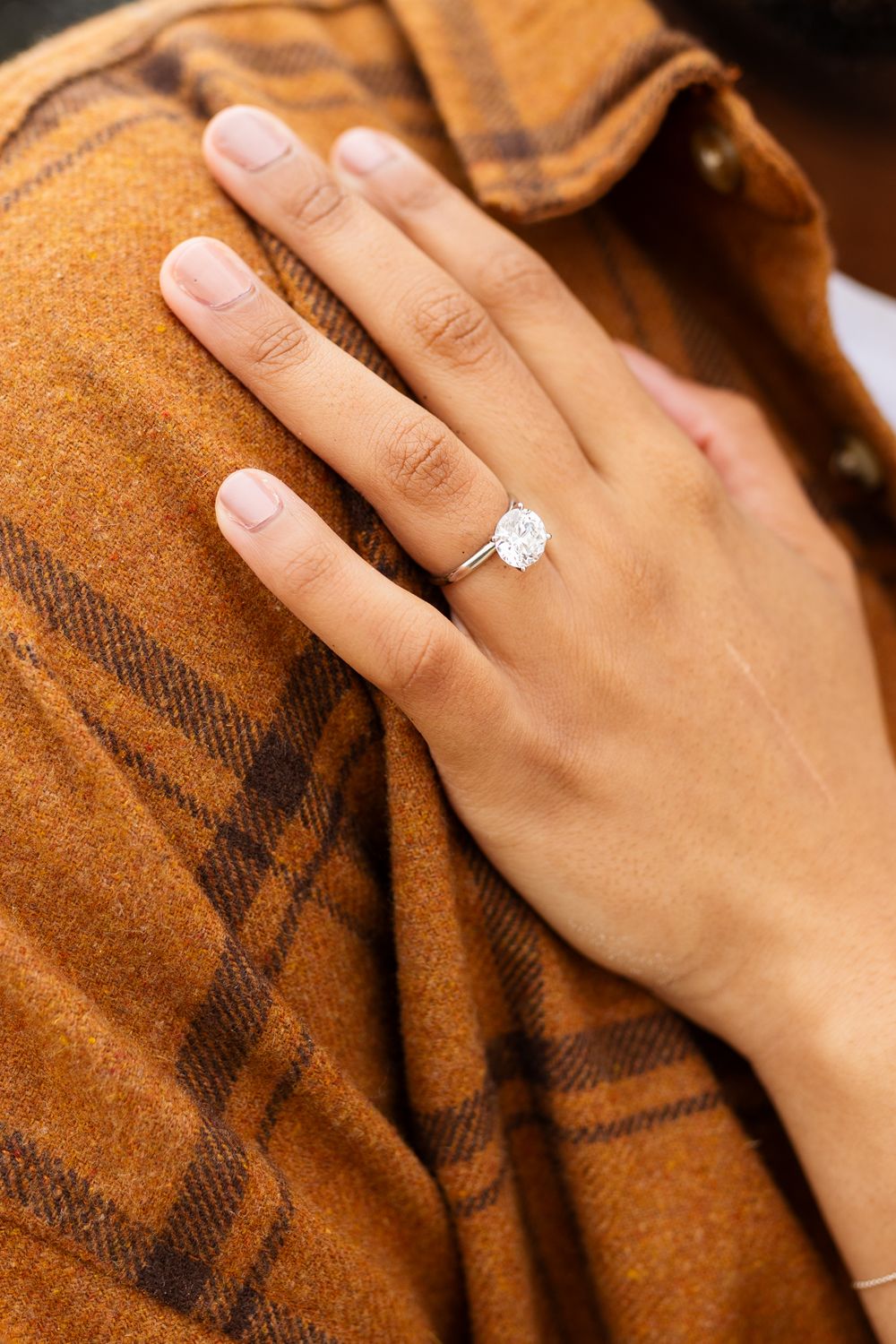 Close-up of a hand wearing a solitaire diamond engagement ring on a ring finger, resting on a brown plaid jacket.