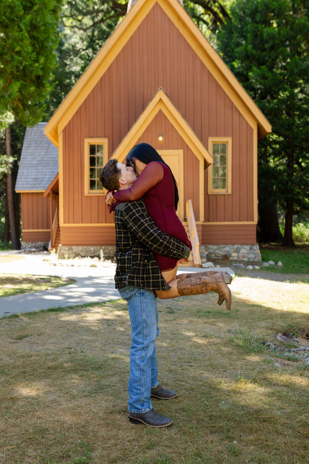 Couple sharing a romantic kiss, man lifting woman in front of a charming wooden chapel surrounded by trees.