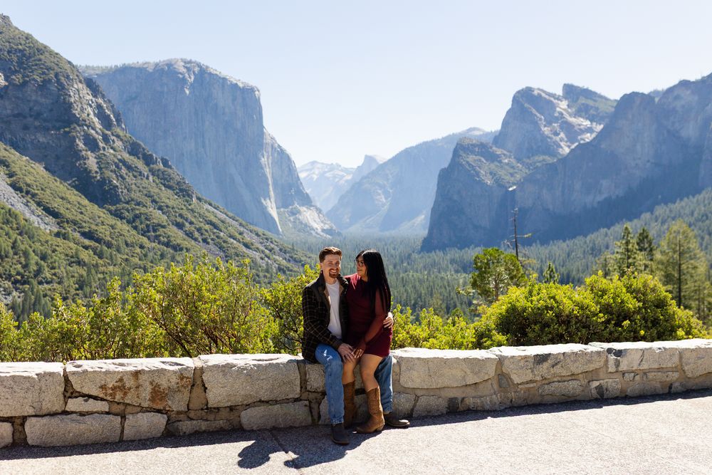 Couple sitting on stone wall at Yosemite Valley tunnel view overlook with mountains and waterfall backdrop.