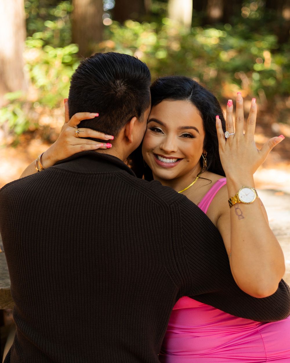 A joyful woman in a pink dress shows off her engagement ring while embracing her partner outdoors in a wooded setting.