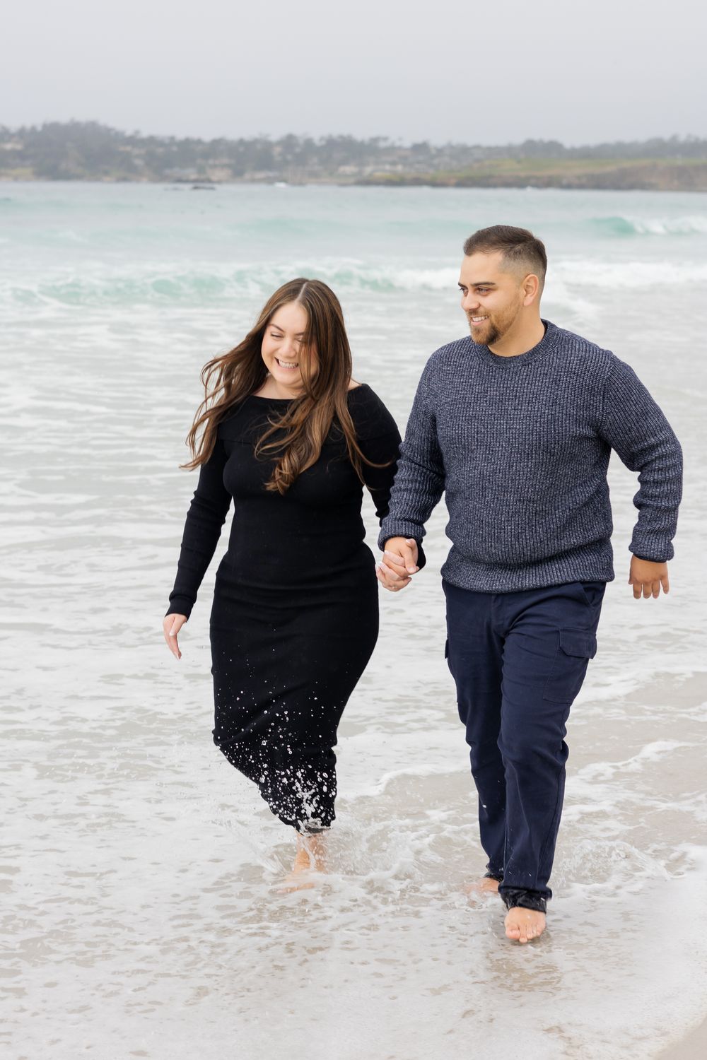 Couple holding hands walking barefoot on a sandy beach, woman in black dress and man in navy sweater and pants.