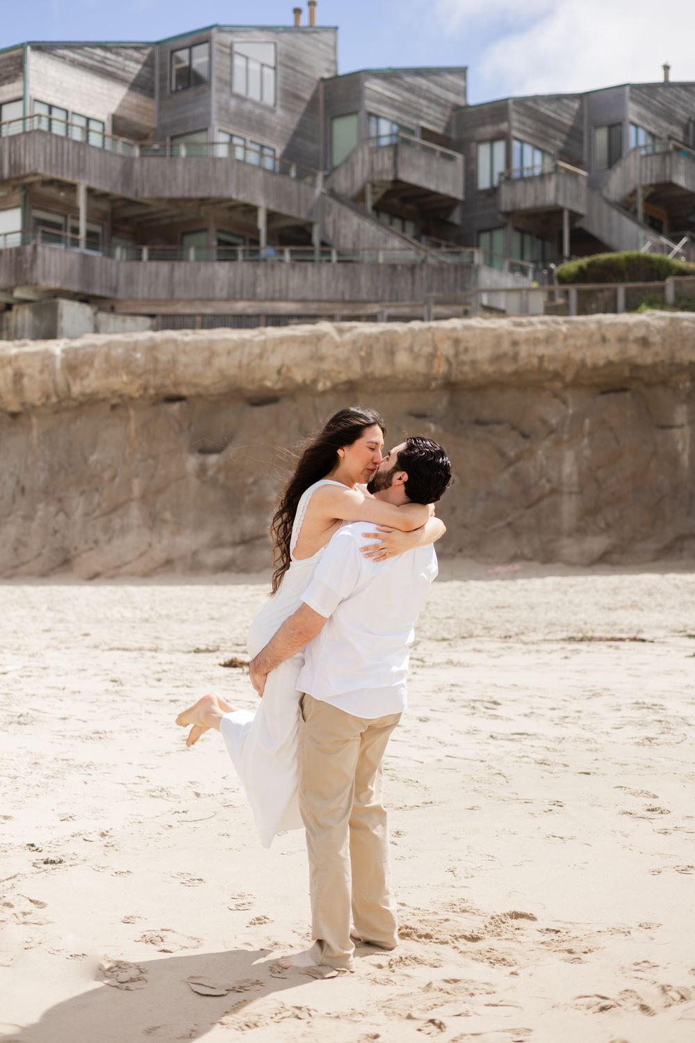 Couple sharing a romantic kiss on a sandy beach, man lifting woman in white dress, with coastal building in background.