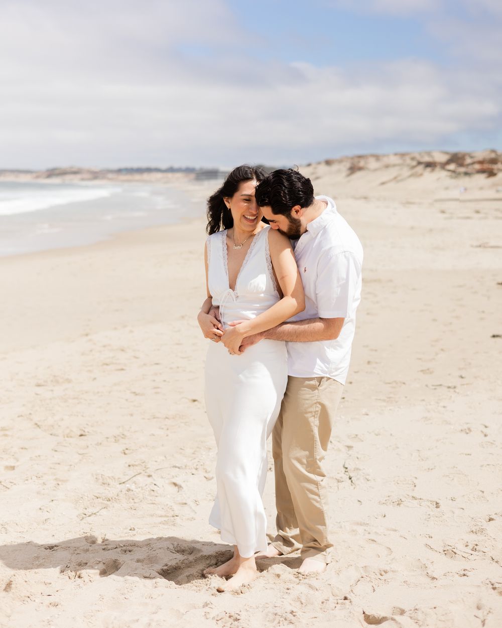 A couple embraces on a sandy beach, the woman in a white dress smiling as the man kisses her cheek.