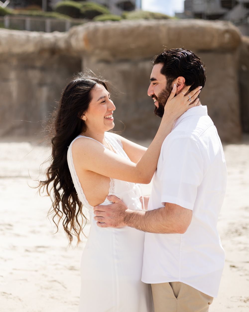 Couple in white outfits sharing a romantic moment on a sandy beach, smiling and embracing each other.