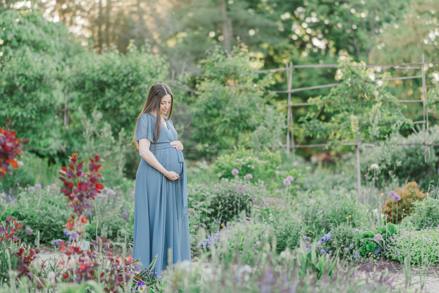 Pregnant woman in blue maxi dress standing in lush garden, cradling baby bump during outdoor maternity photo session.