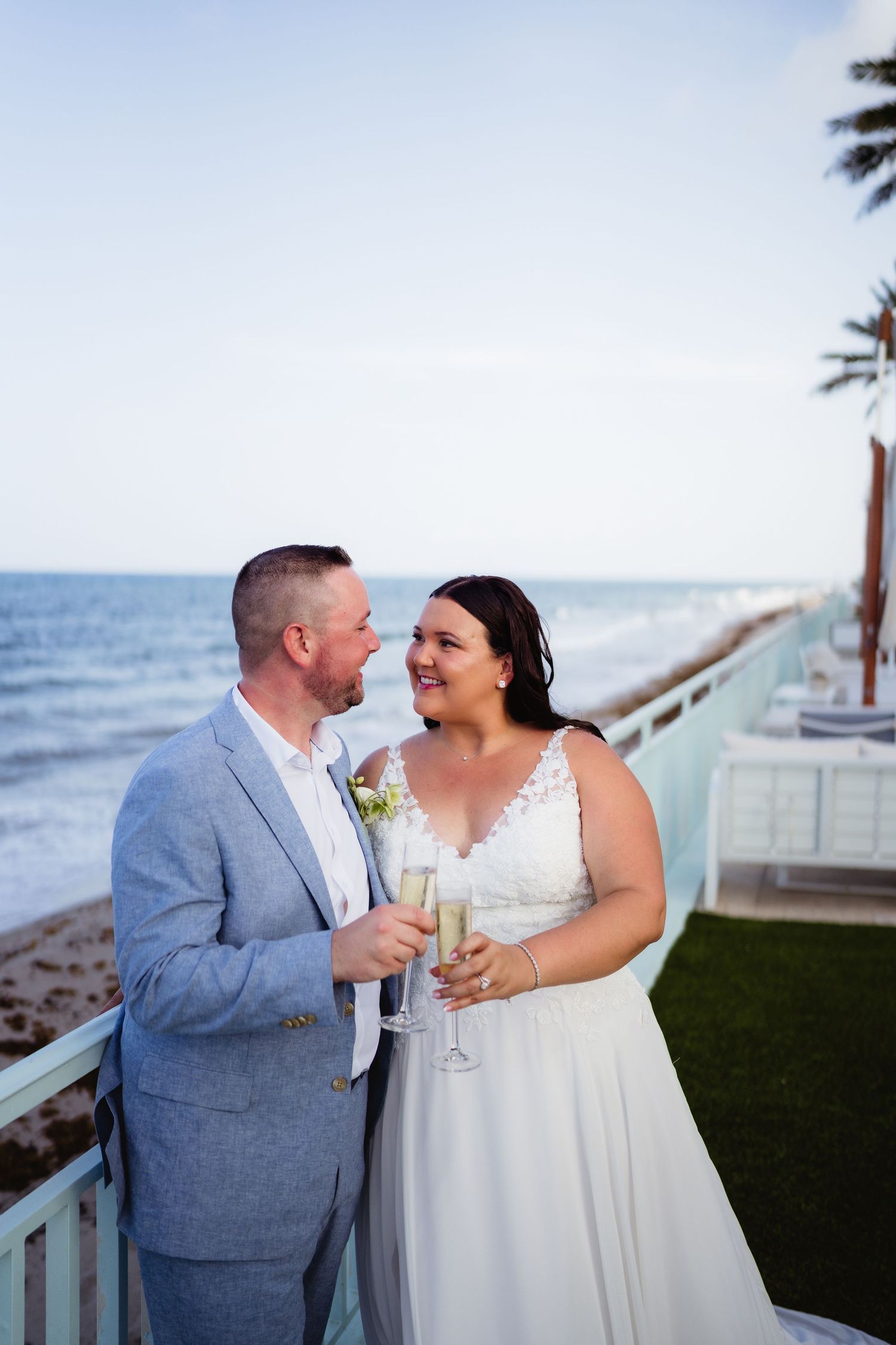 Newlywed couple toasting with champagne on a beachfront balcony, bride in white gown and groom in light blue suit.
