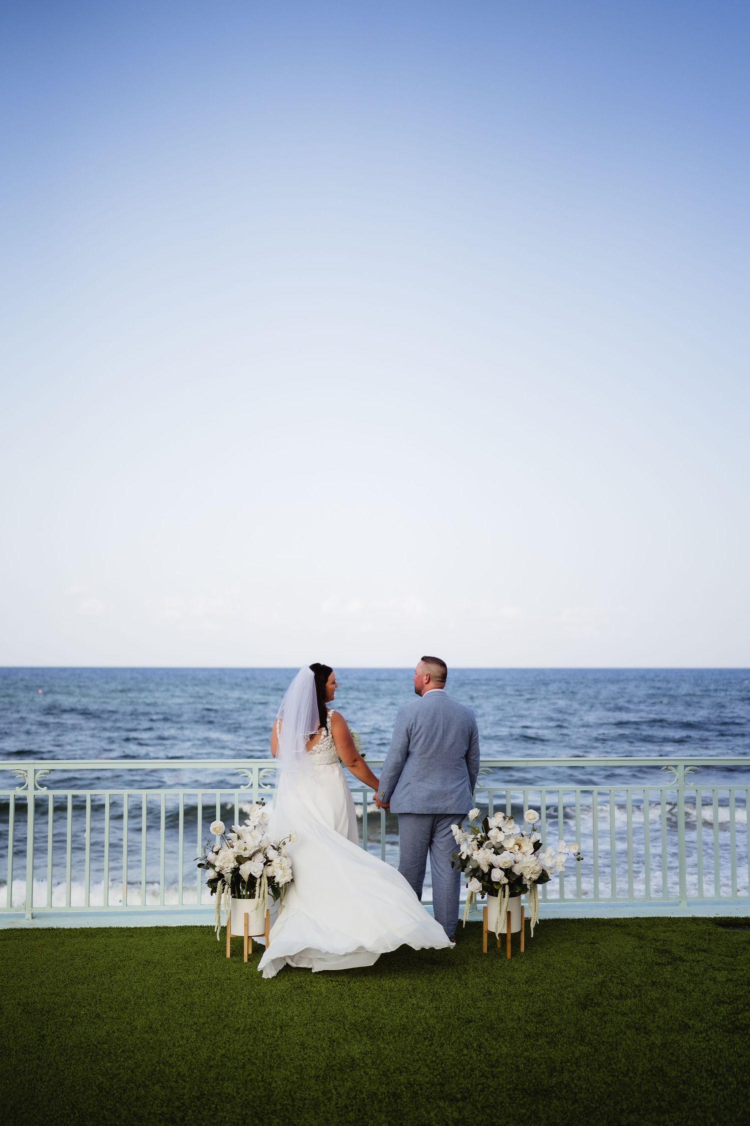Bride and groom stand hand in hand at oceanfront wedding ceremony with white floral arrangements and railing backdrop.