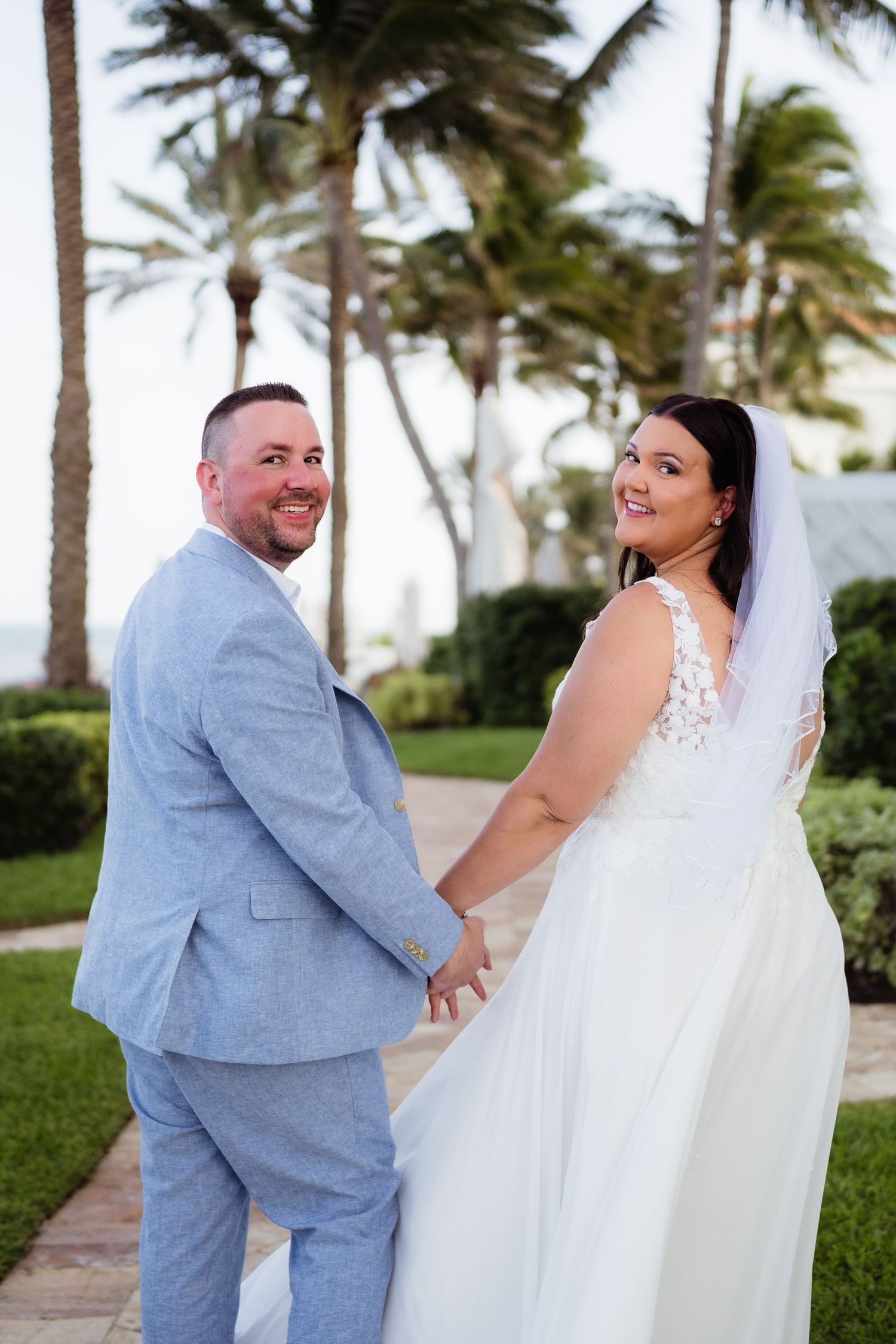 Bride and groom holding hands smiling at a tropical outdoor wedding venue with palm trees.