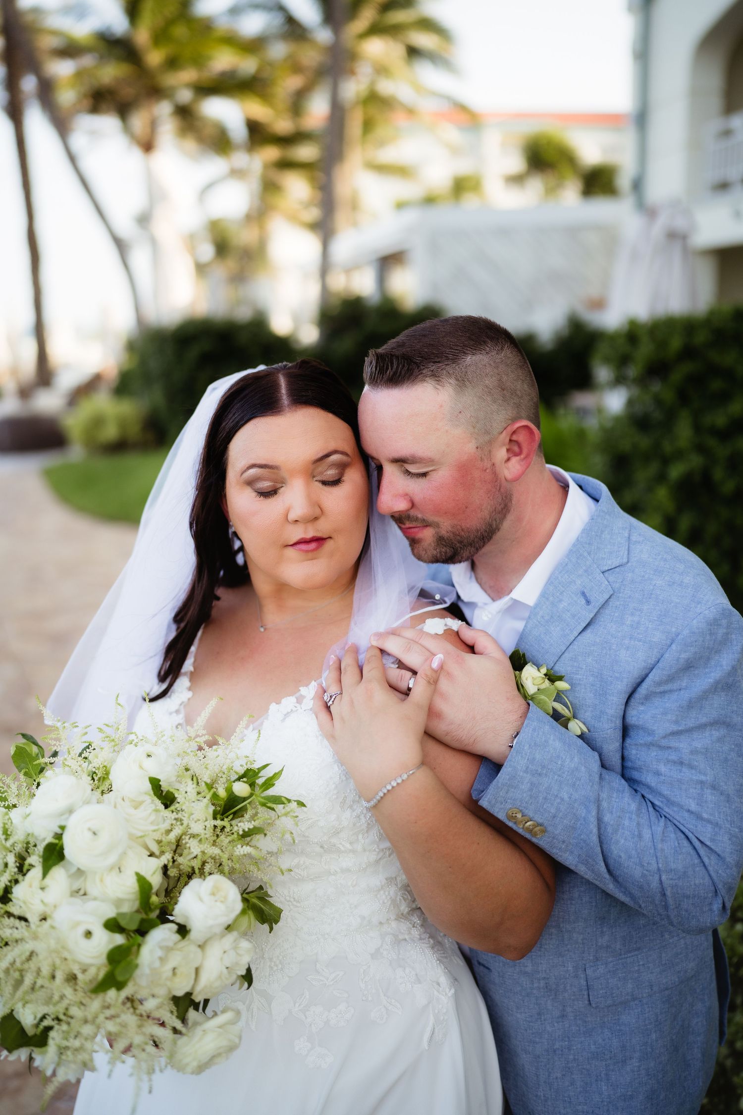 Bride and groom share a tender embrace outdoors at a tropical wedding venue, bride holding white floral bouquet.