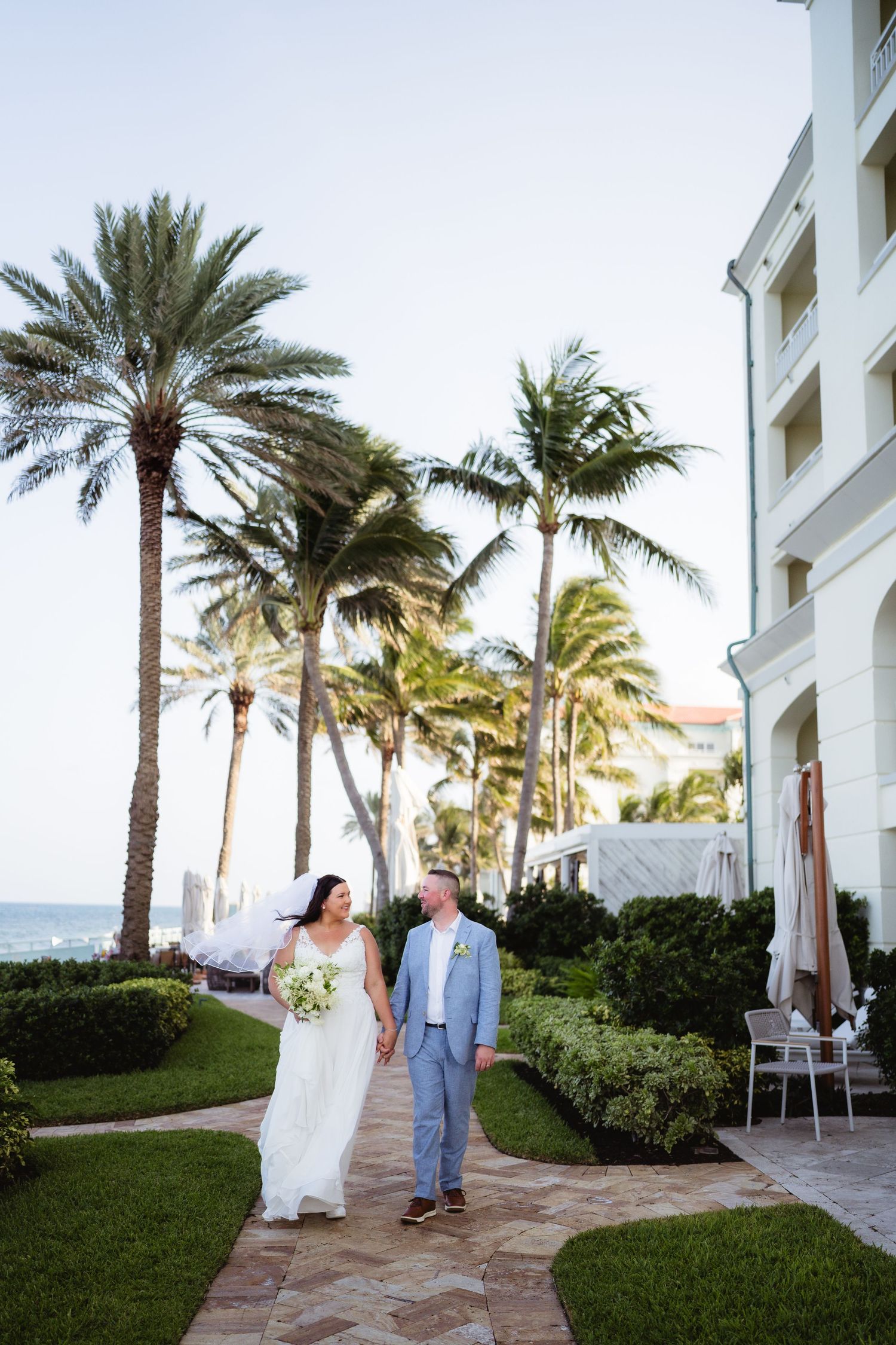 Bride and groom walk hand-in-hand along a tropical palm-lined path near a beachside resort.