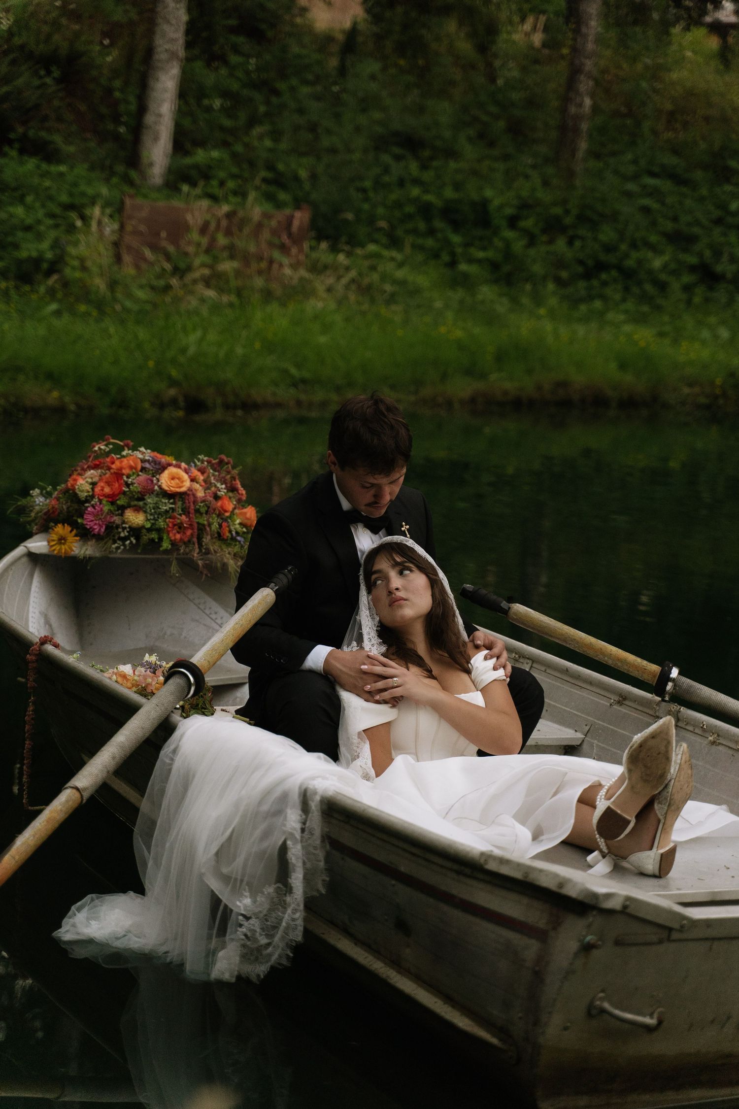 Bride and groom in wedding attire share a romantic moment on a flower-adorned rowboat on a serene lake.
