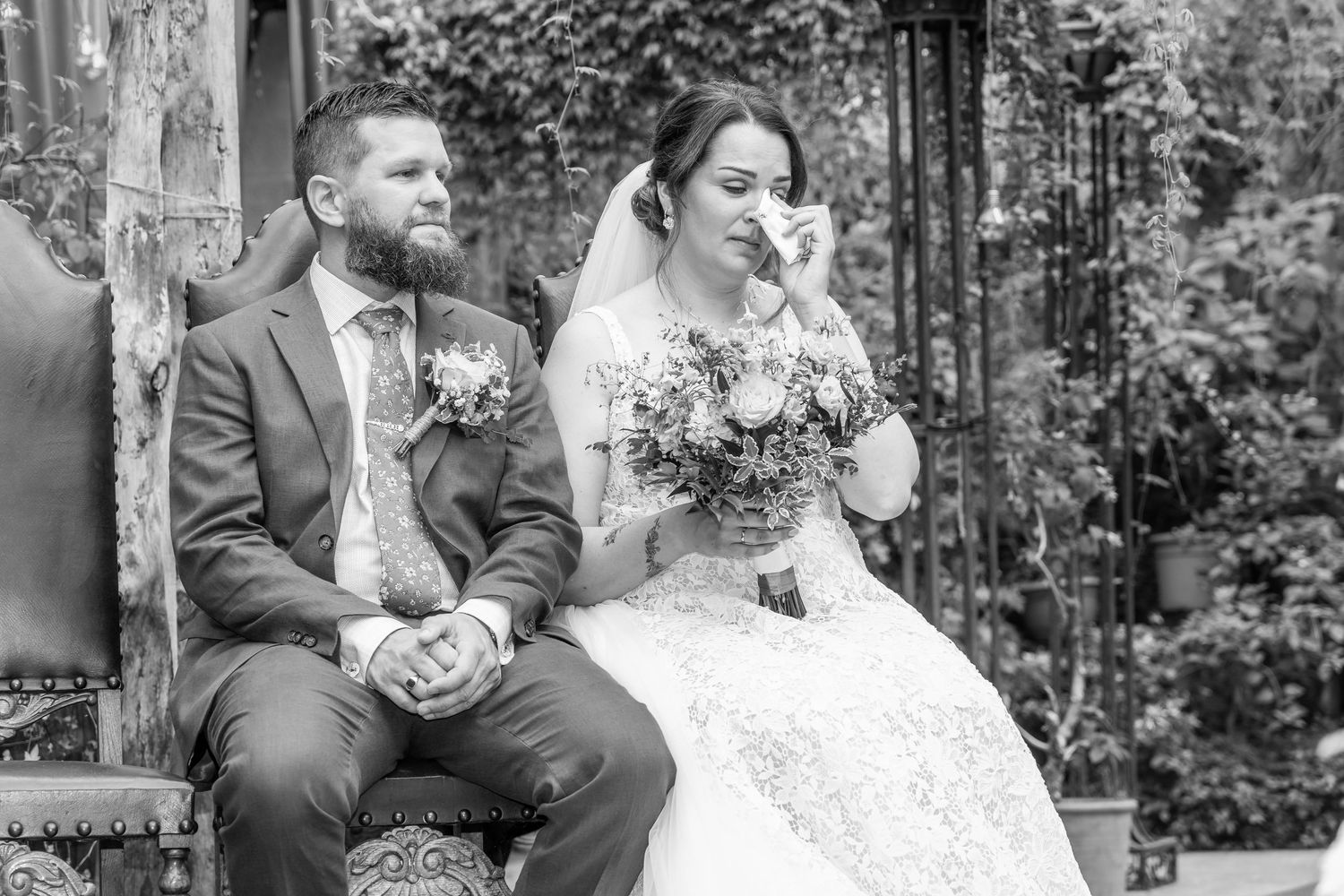 Emotional bride dabs tears during wedding ceremony while groom sits beside her in black and white photo.