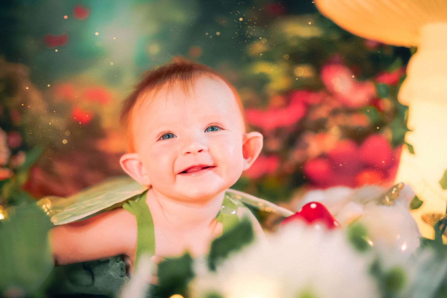 Smiling baby in green outfit surrounded by colorful flowers and bokeh lights in a whimsical garden setting a home session in Lake Worth, FL.