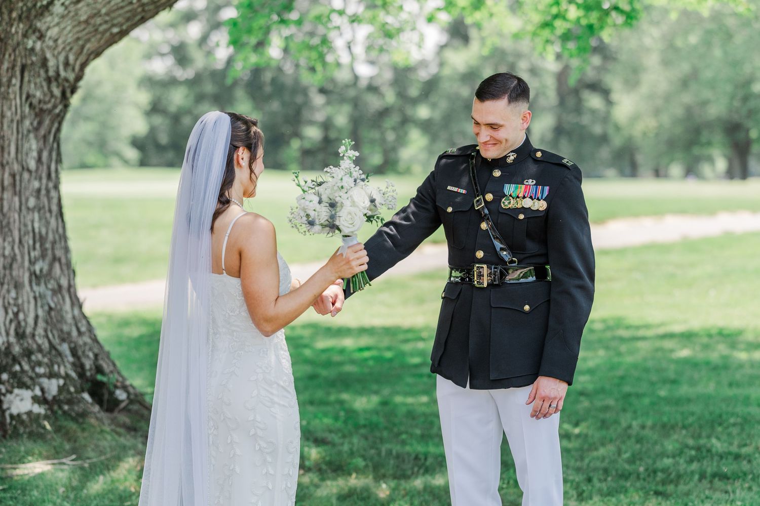 Marine groom in dress uniform holds bride's hand outdoors under tree at military wedding ceremony.
