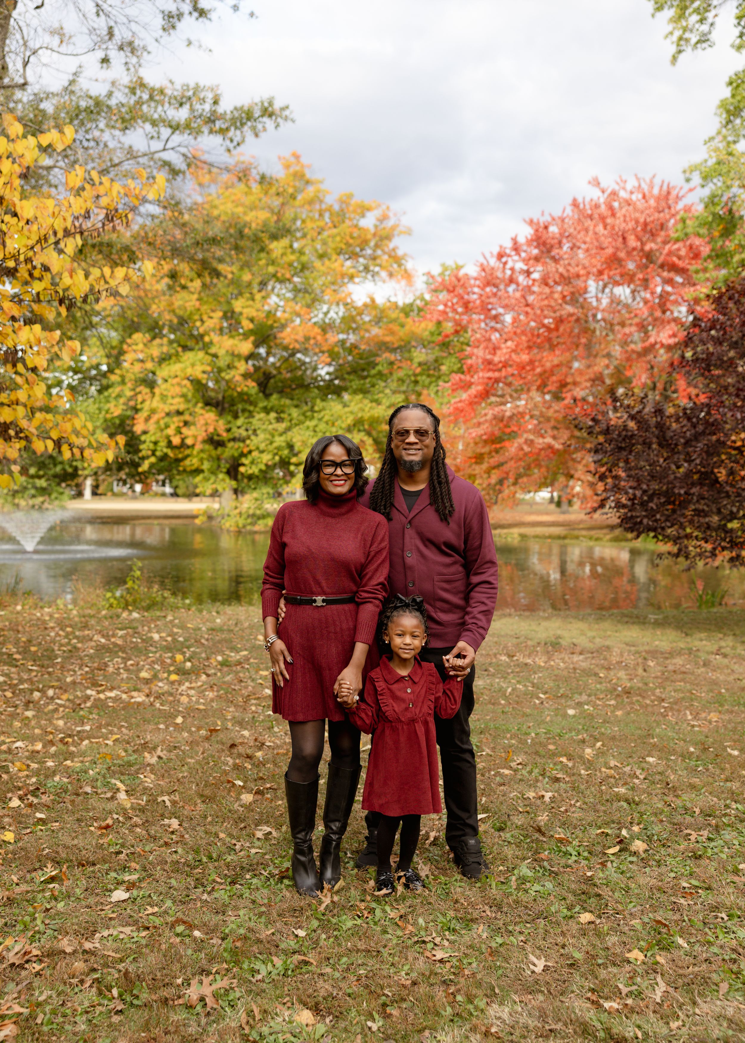 Family of three in matching burgundy outfits posing outdoors among vibrant fall foliage near a pond.