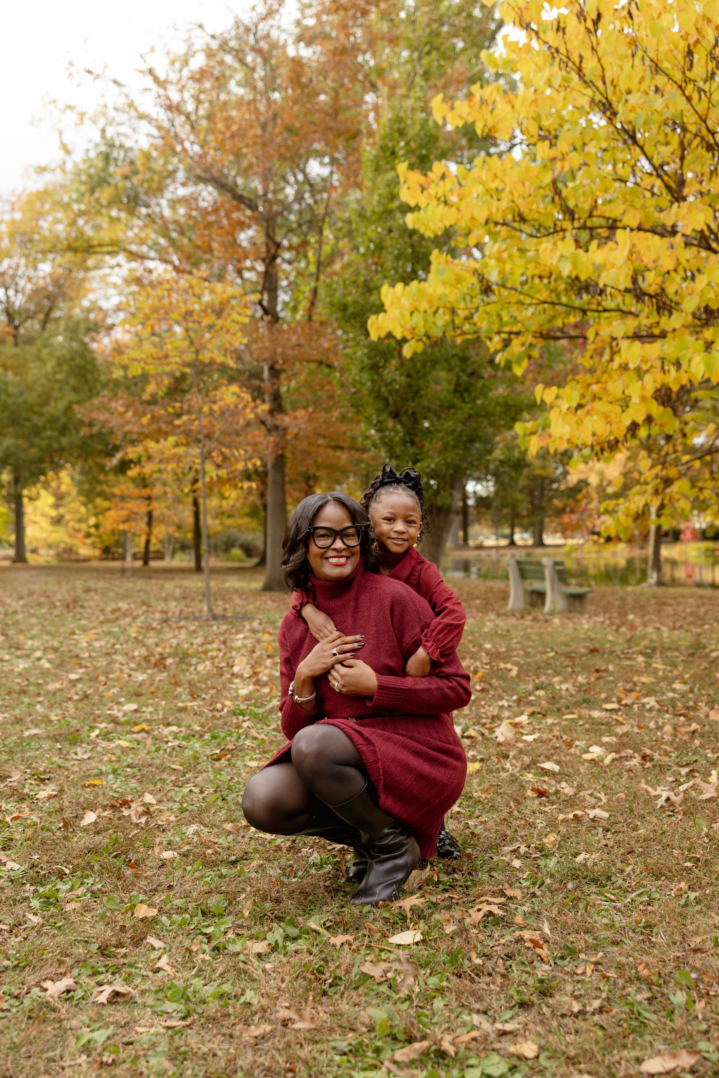 Mother and daughter in matching burgundy dresses posing in autumn park with colorful yellow foliage.