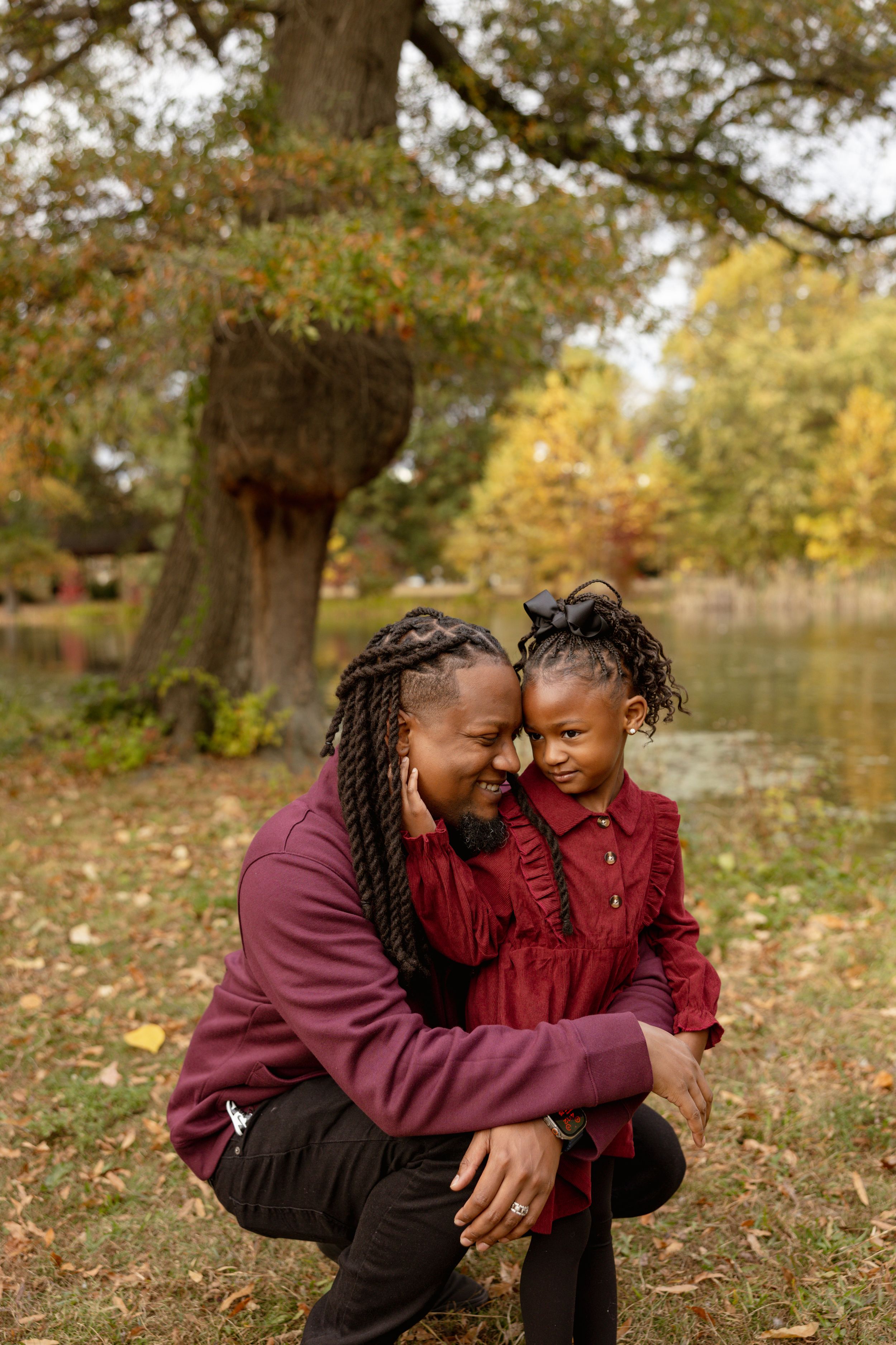 A mother and young daughter in matching burgundy outfits share a tender forehead touch in an autumn park.