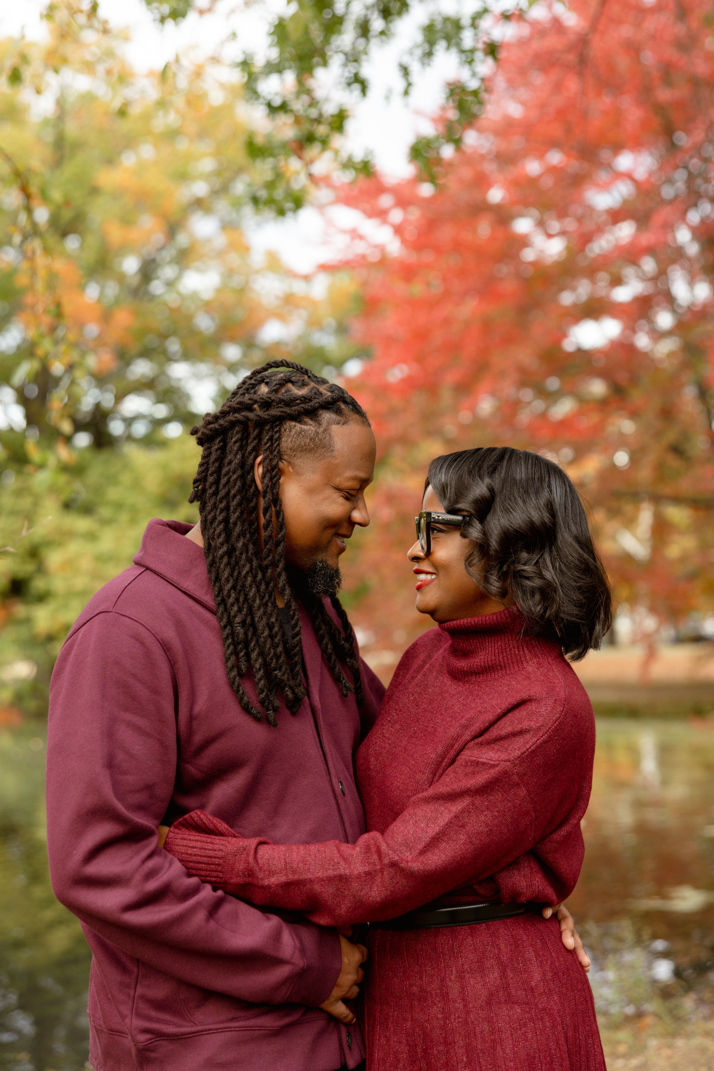 A couple embraces and smiles at each other outdoors, surrounded by vibrant autumn foliage near a pond.