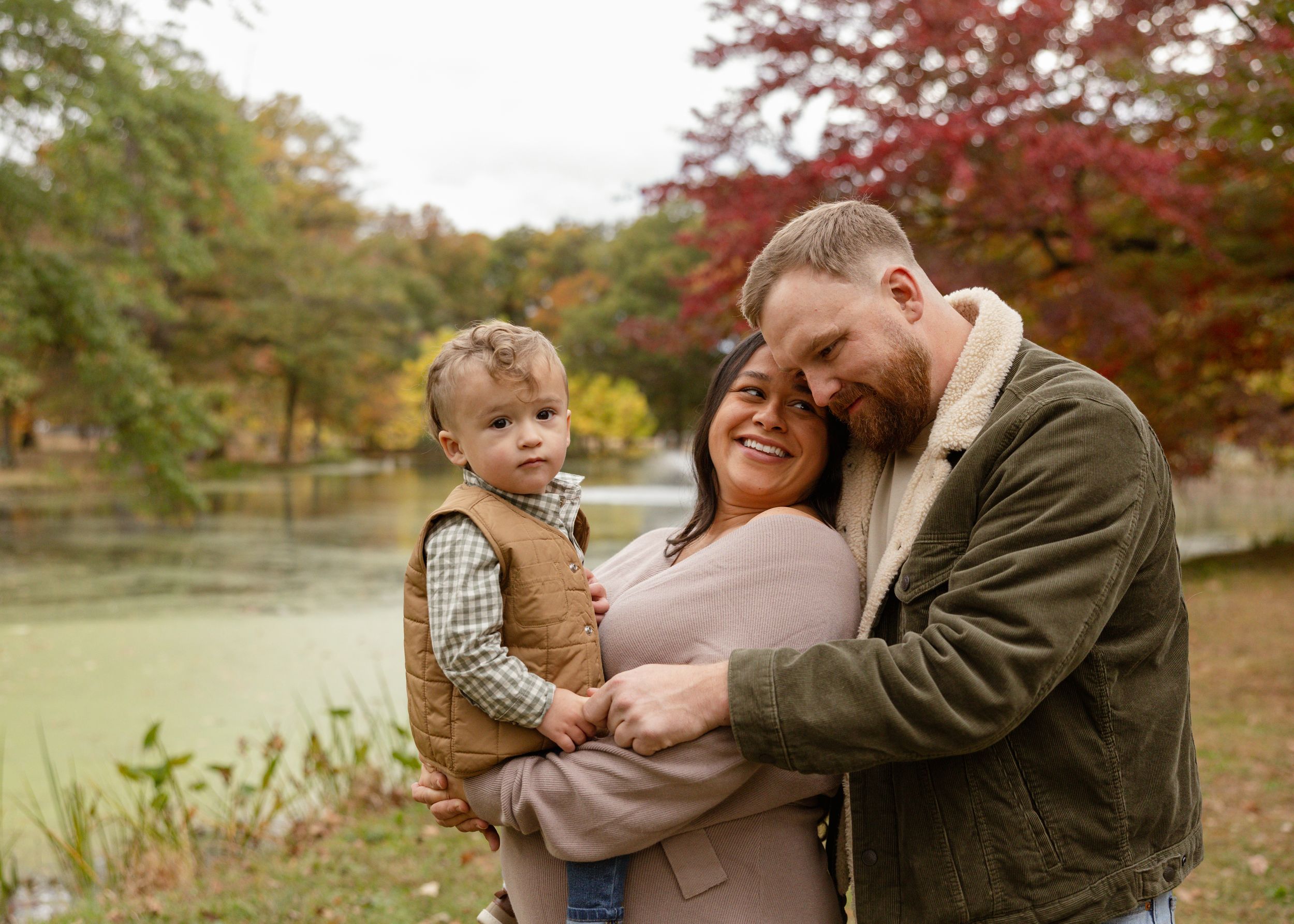 Family of three posing outdoors in autumn park near pond, parents holding toddler son, colorful fall foliage background.
