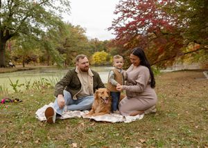 Family with toddler and golden retriever enjoying autumn picnic by pond with colorful fall foliage.