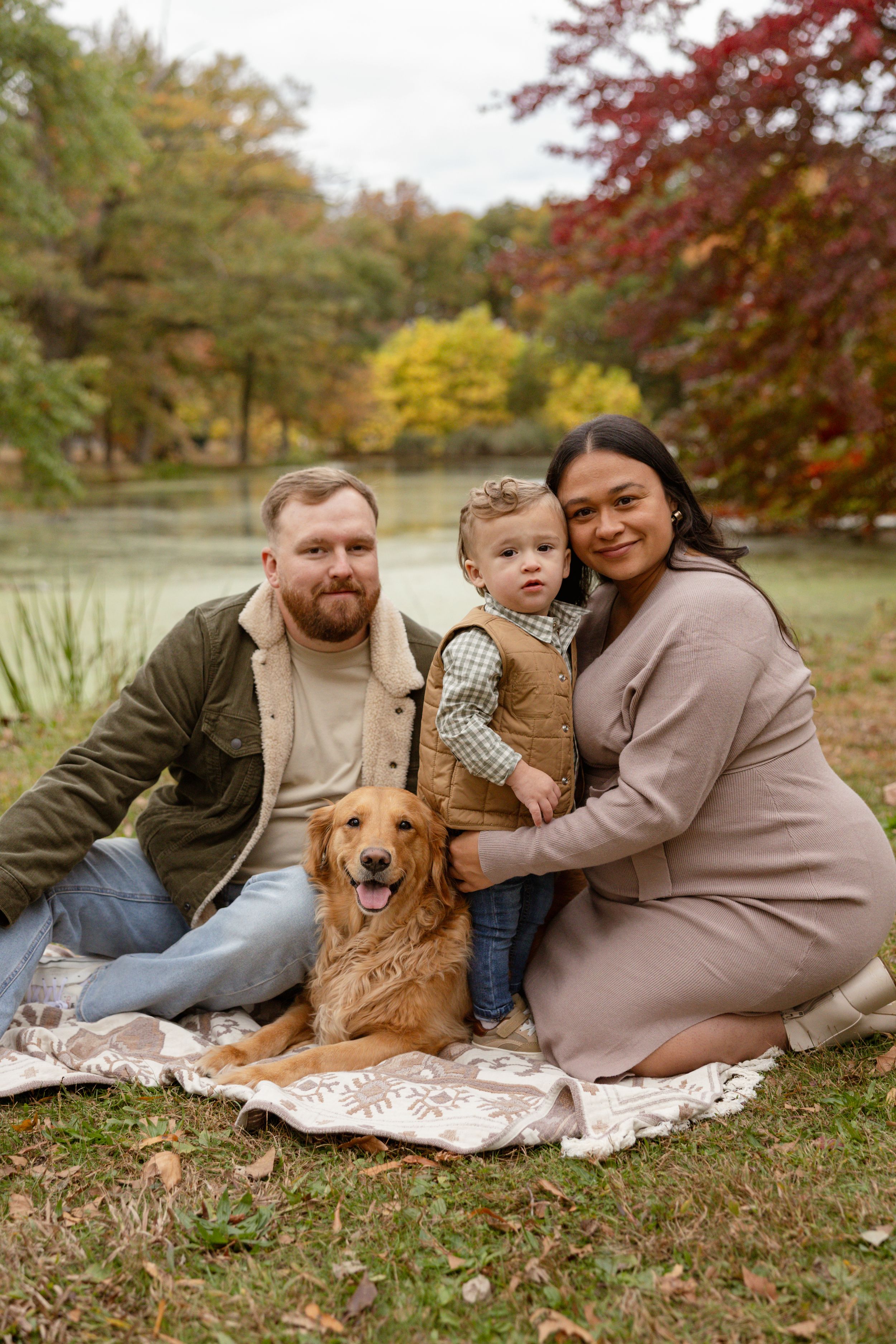 Family with toddler and golden retriever sitting on blanket in autumn park near pond.