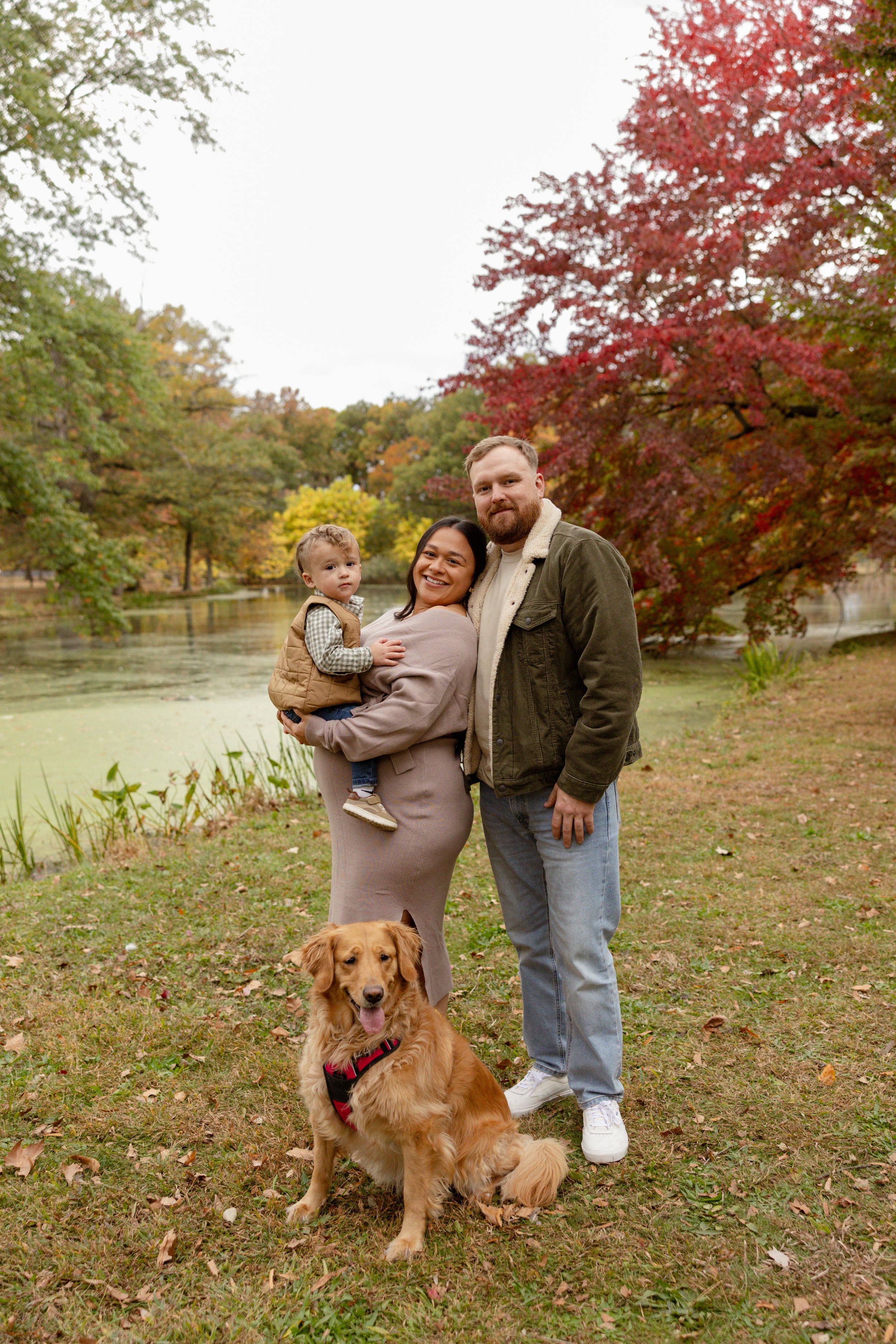Family with toddler and golden retriever posing by a pond surrounded by colorful autumn foliage.