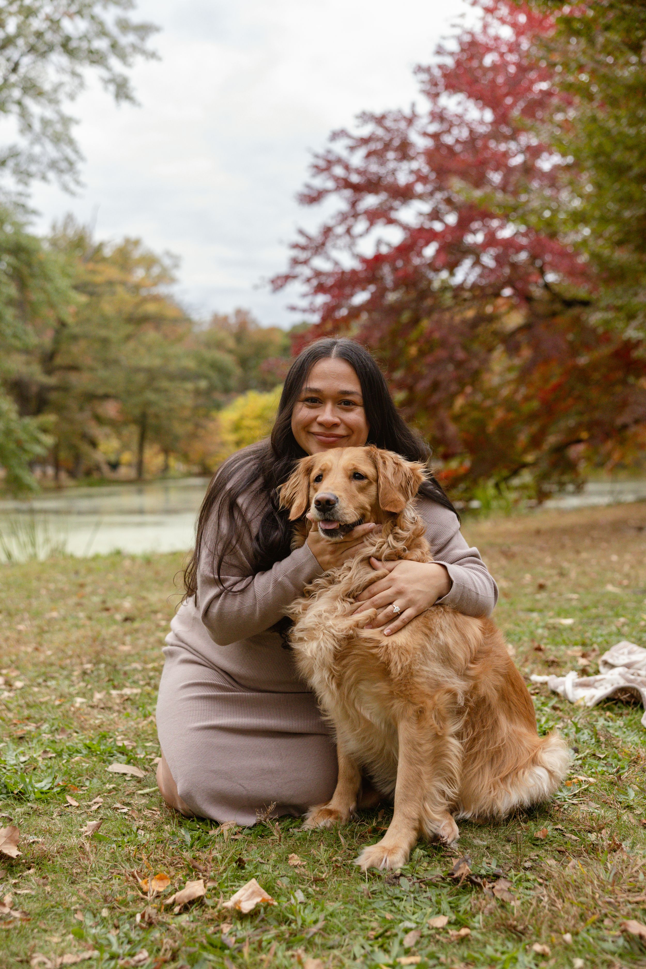 Woman smiling and hugging a golden retriever dog in an autumn park with colorful foliage and a pond in the background.