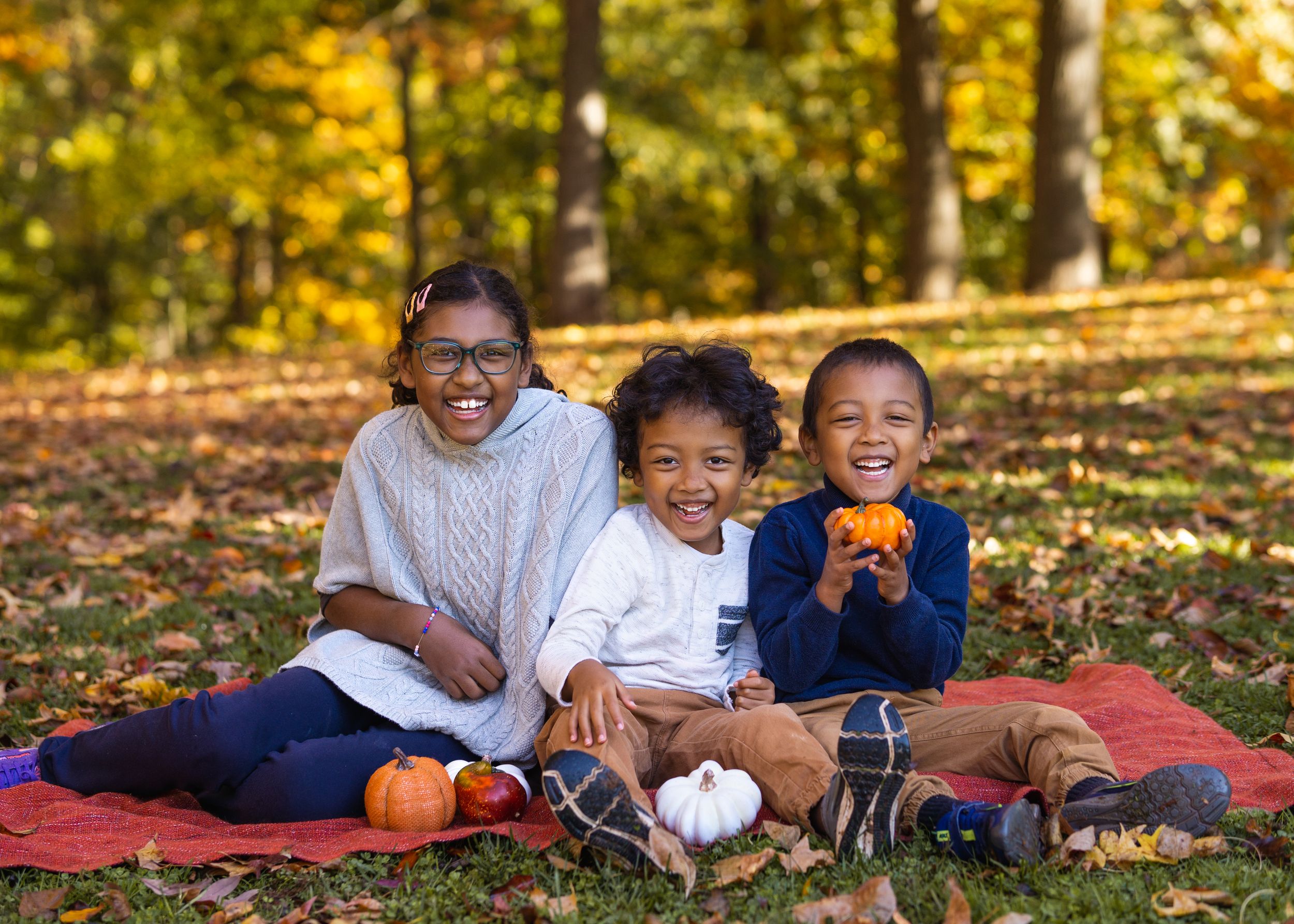 Three children smiling on a red blanket in an autumn park, surrounded by small pumpkins and colorful fall foliage.