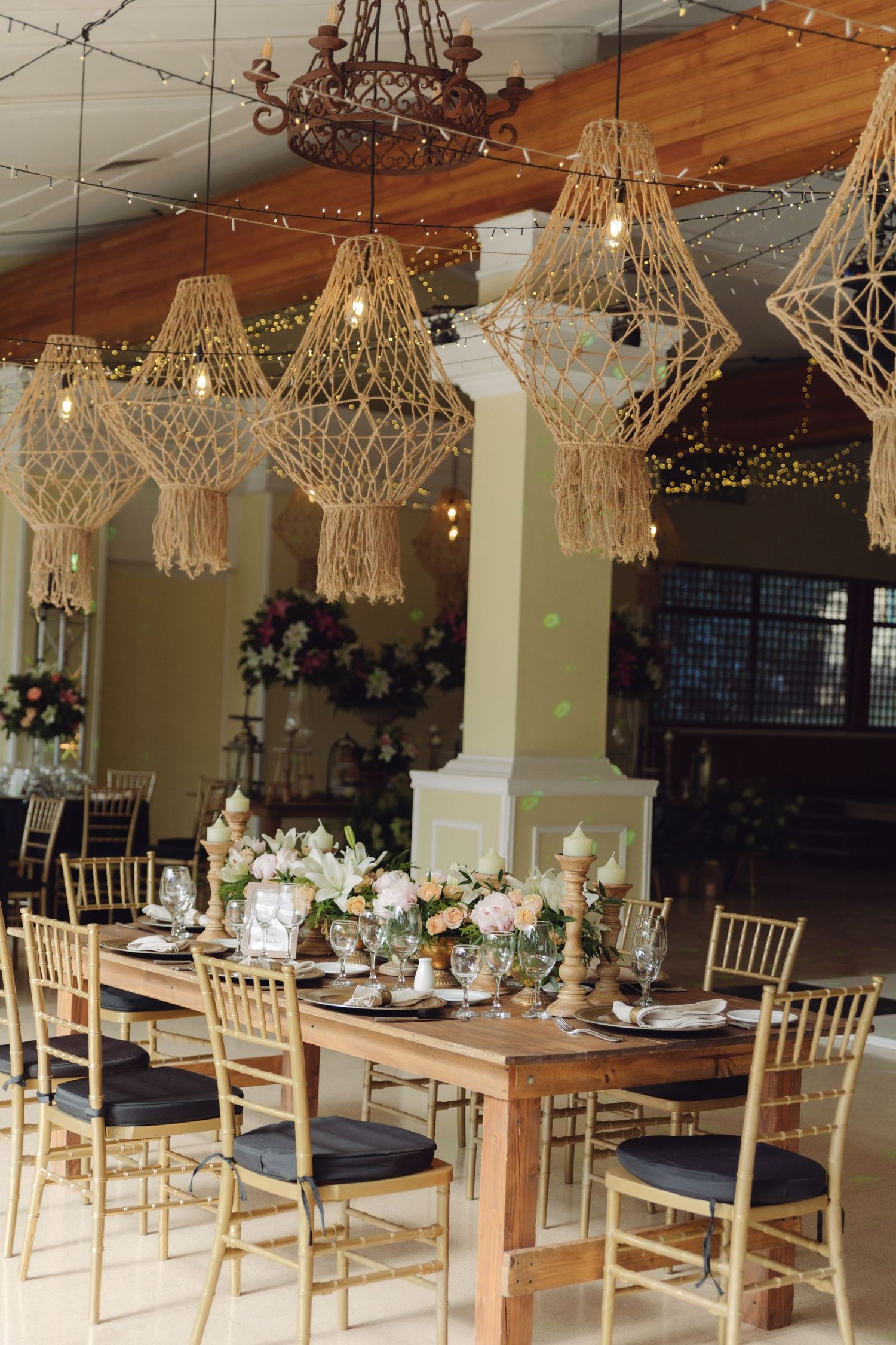 Elegant wedding reception with macrame chandeliers, gold chiavari chairs, and floral centerpieces on rustic wooden tables.