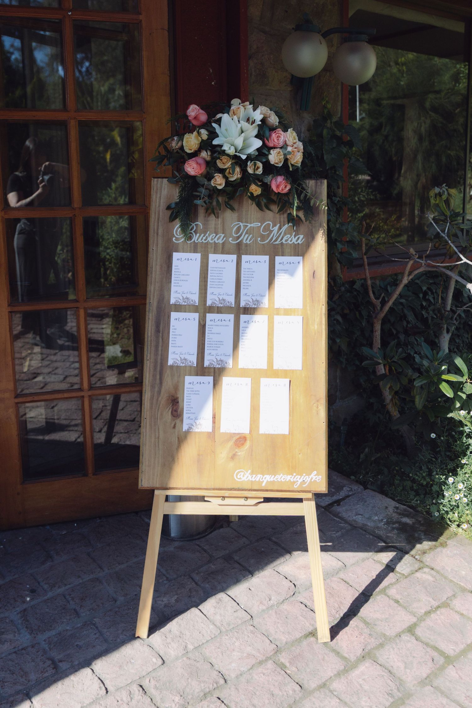 Wooden wedding seating chart easel decorated with pink and white flowers displayed outdoors at venue entrance.