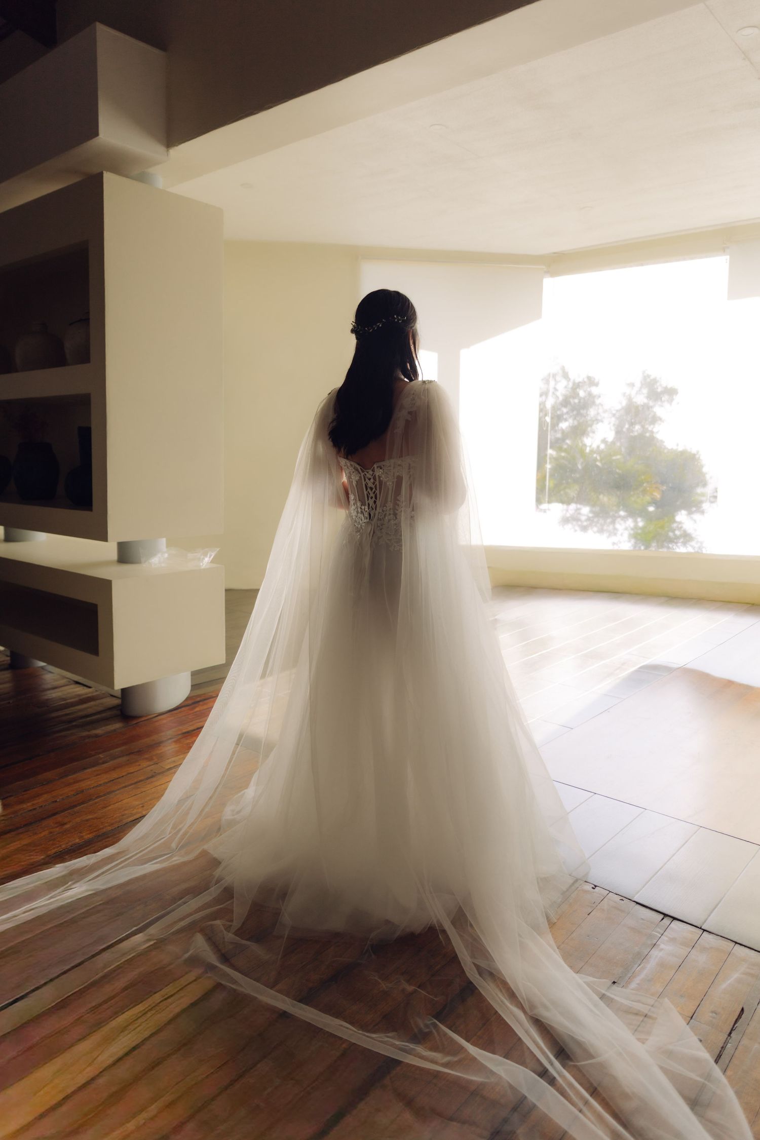 Bride in elegant white lace gown with flowing veil stands facing large window in softly lit interior room.