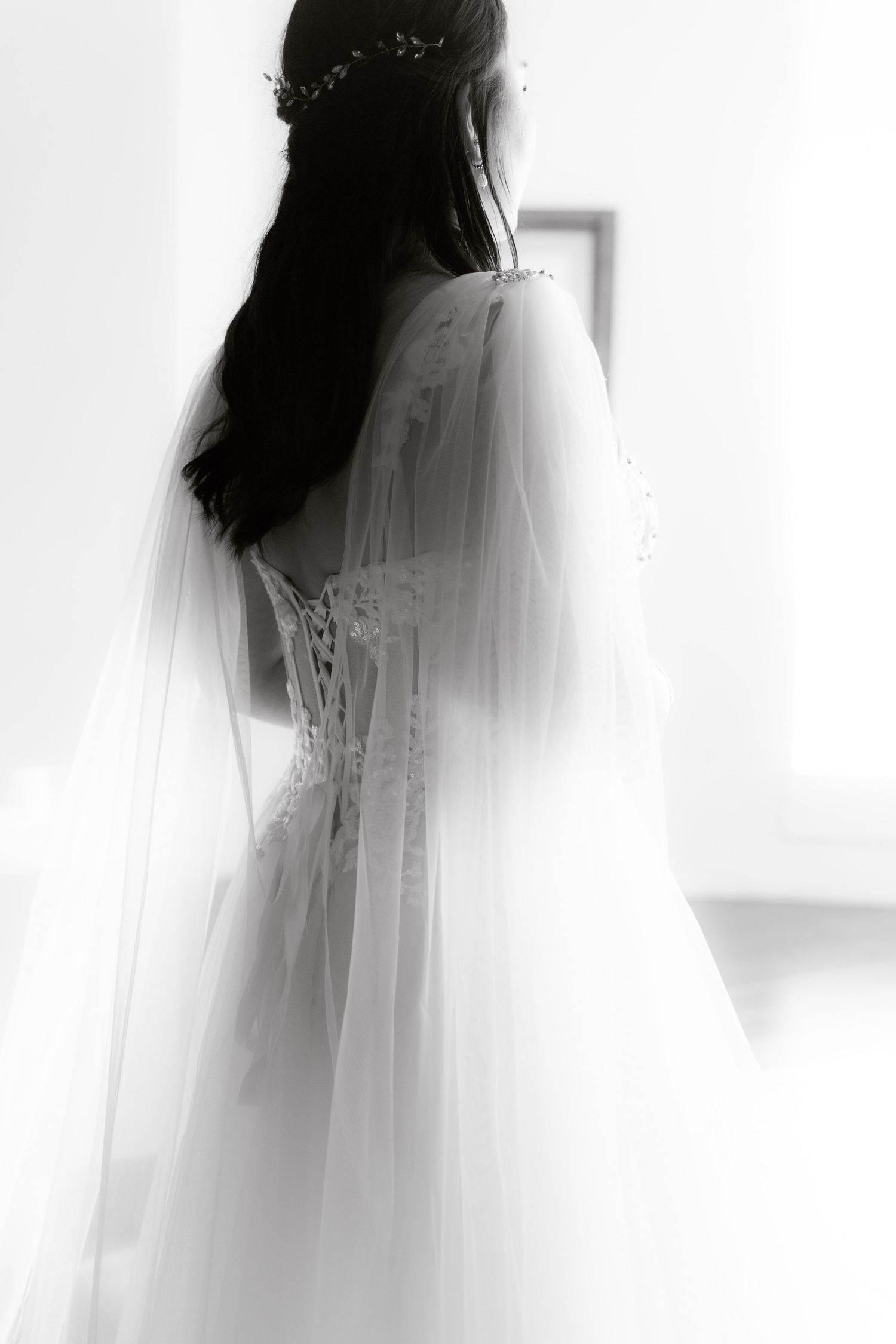 Black and white artistic portrait of bride in lace wedding gown with flowing veil and floral hair crown.