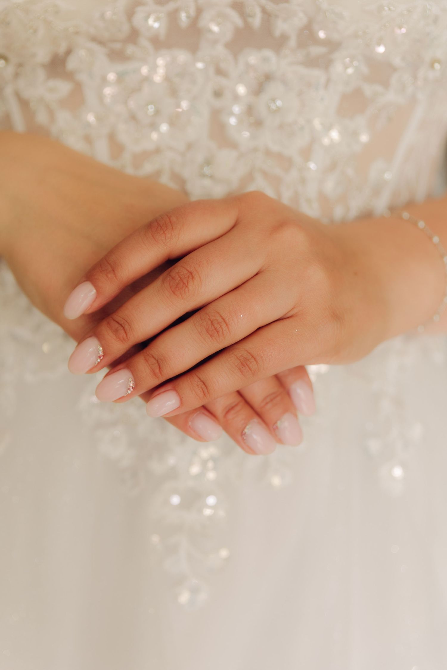 Close-up of brides hands with soft pink manicure clasped together against intricate pearl-embellished wedding dress bodice.