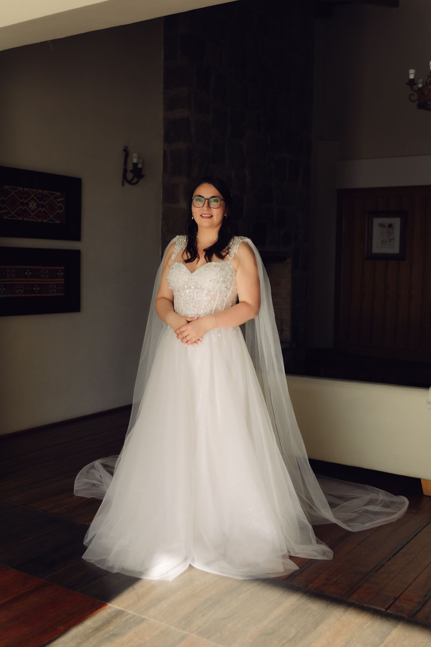 Bride in white lace A-line wedding gown with cape veil stands smiling in elegant indoor setting.