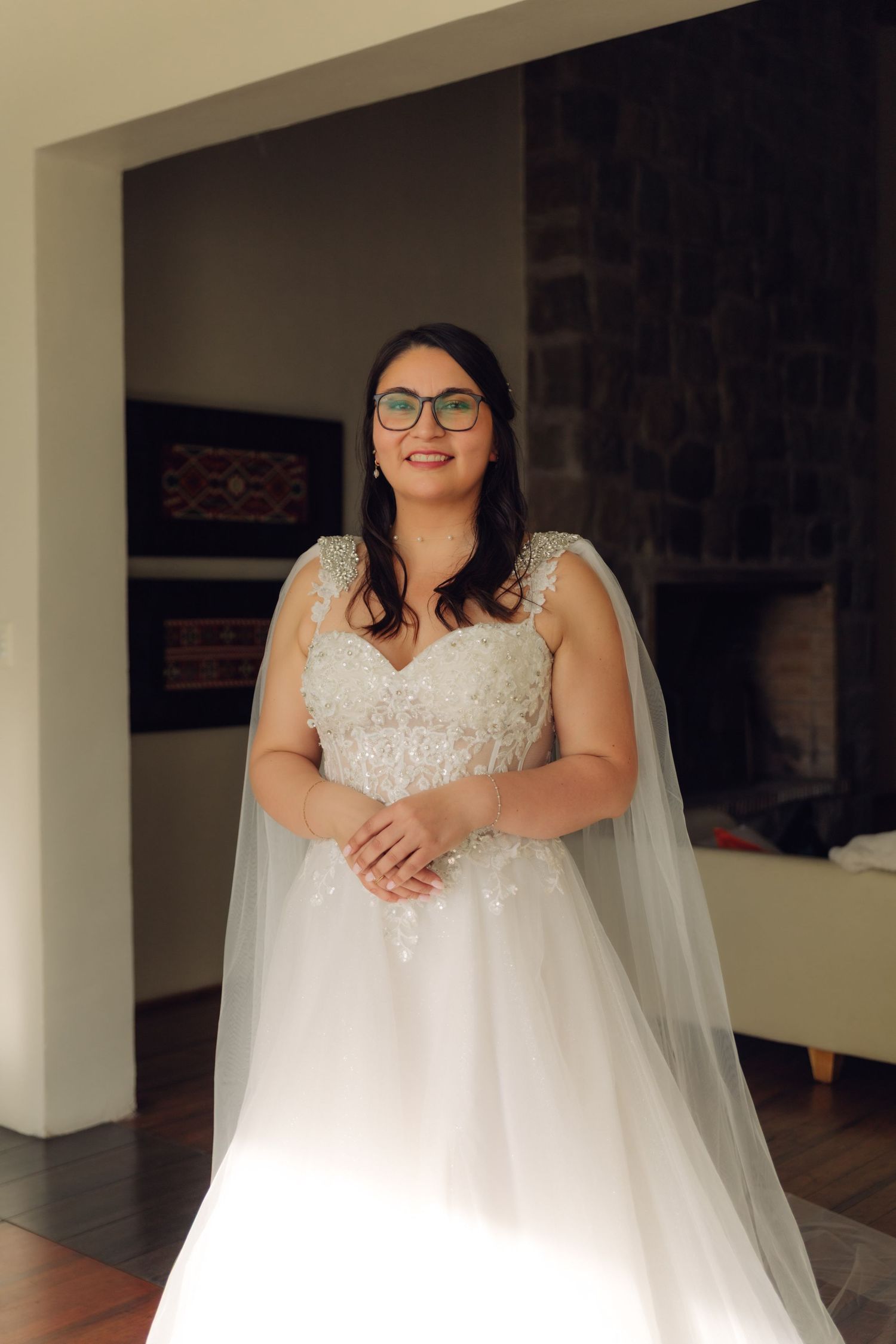 Smiling bride wearing glasses in beaded lace wedding dress with flowing cape veil indoors.