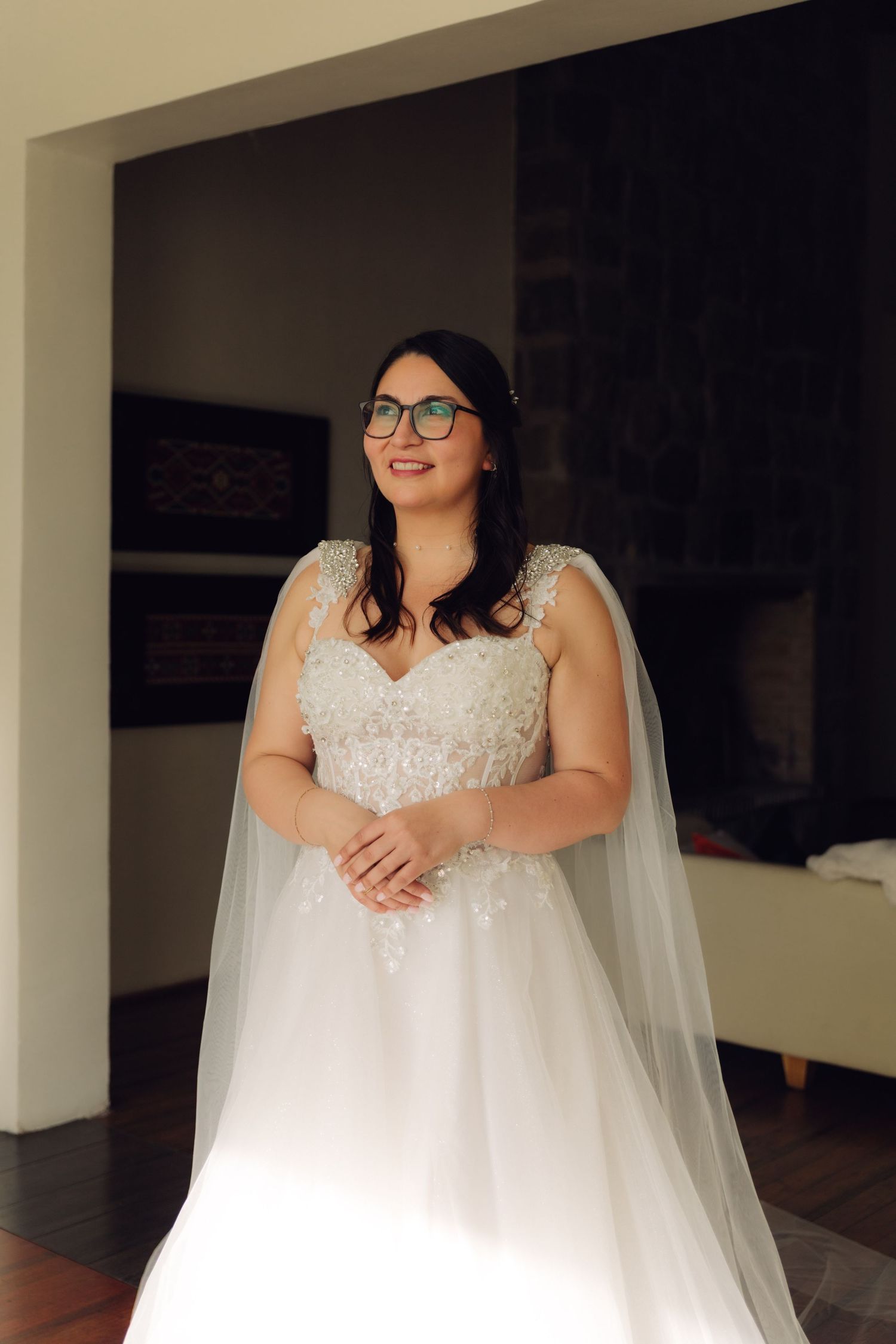 Brunette bride in glasses and white lace wedding gown with cape veil gazes to the side.