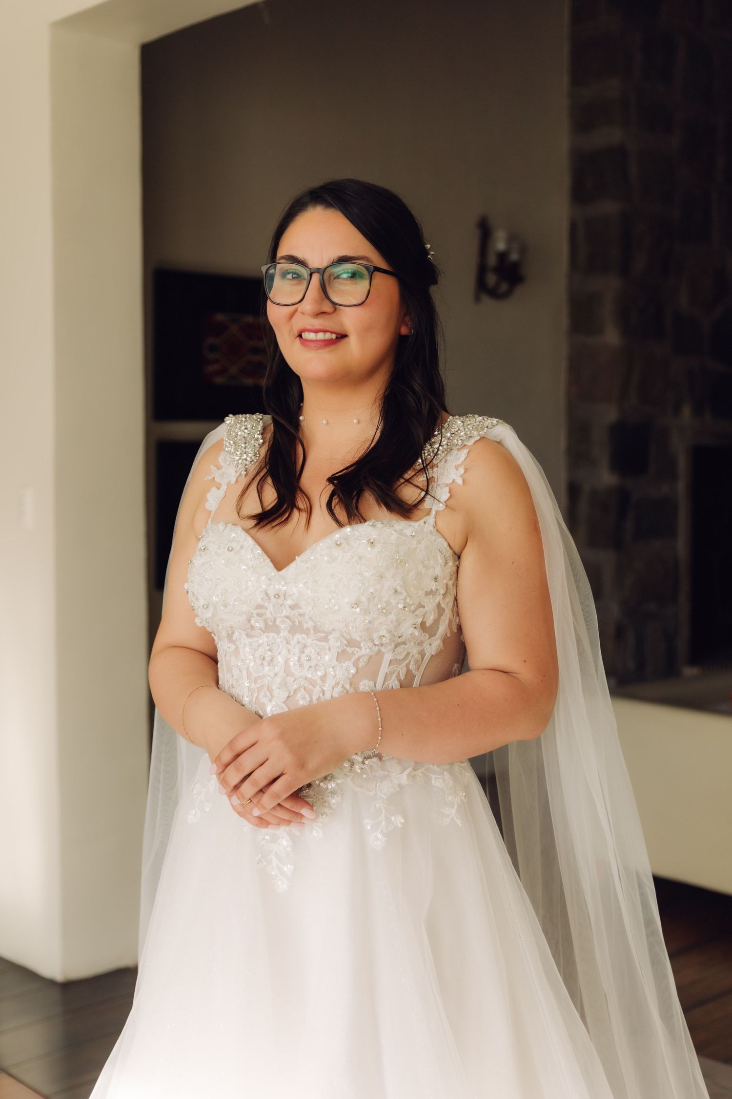 Radiant bride in pearl-embellished lace wedding dress and cape veil smiles in natural light.