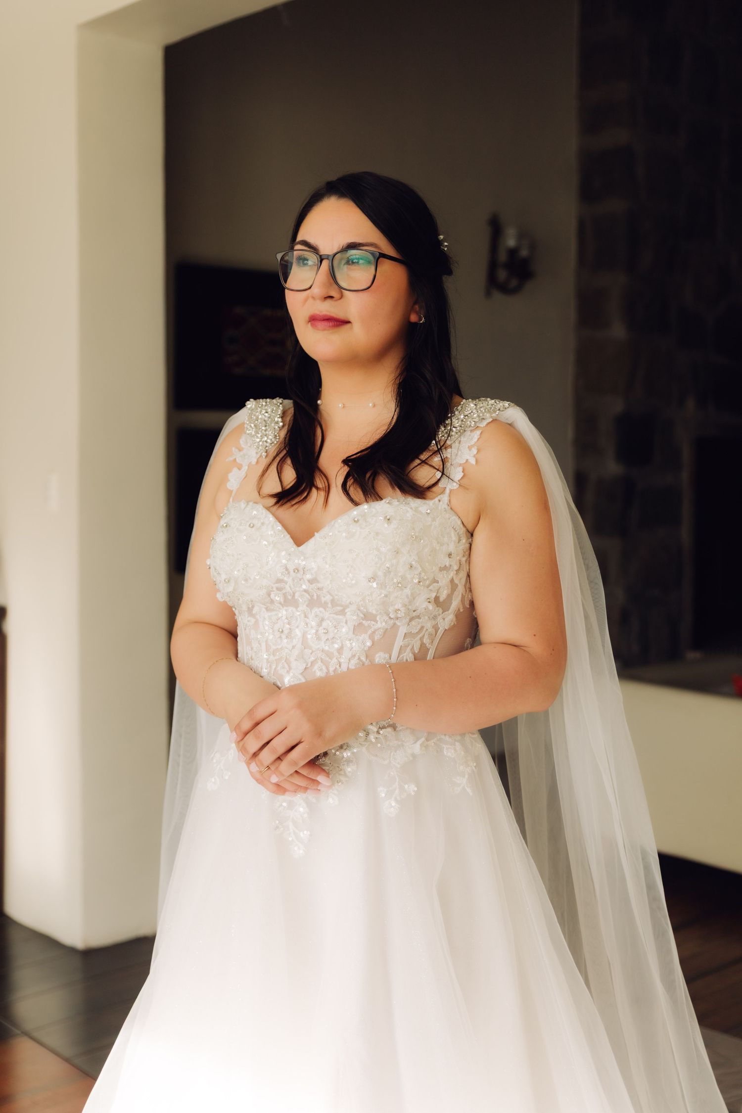 Elegant bride in white lace gown with cape veil and glasses poses in thoughtful expression.