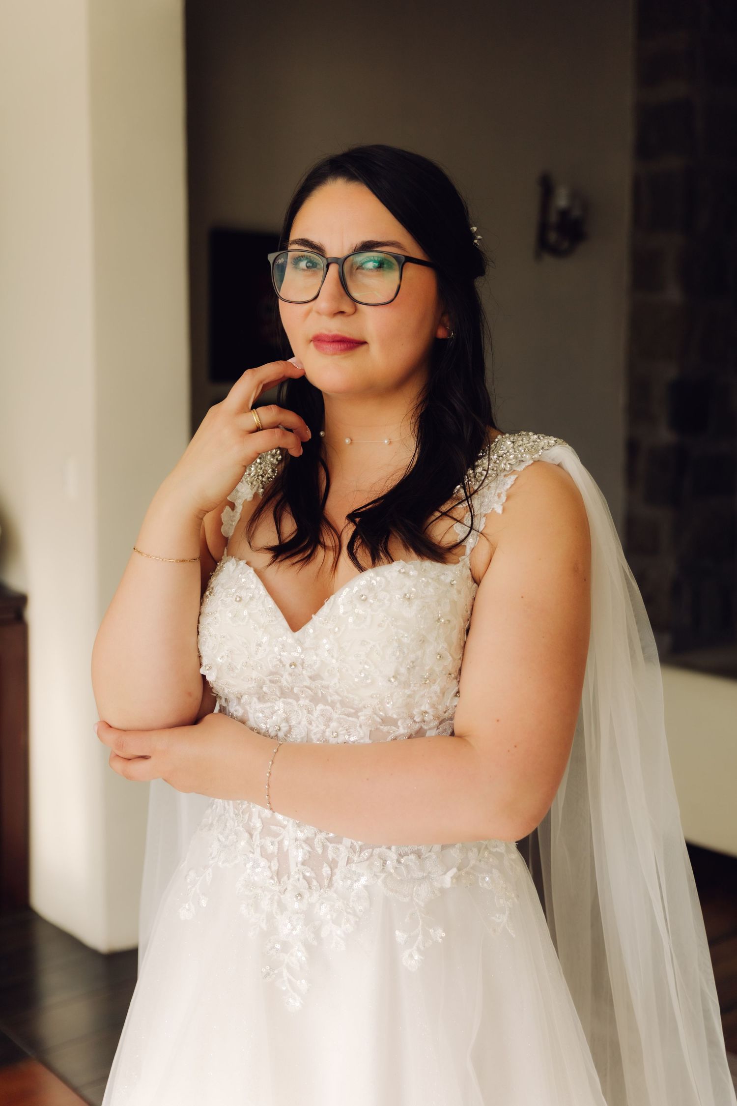 Bride in lace wedding dress and glasses poses with hand near face in contemplative portrait.