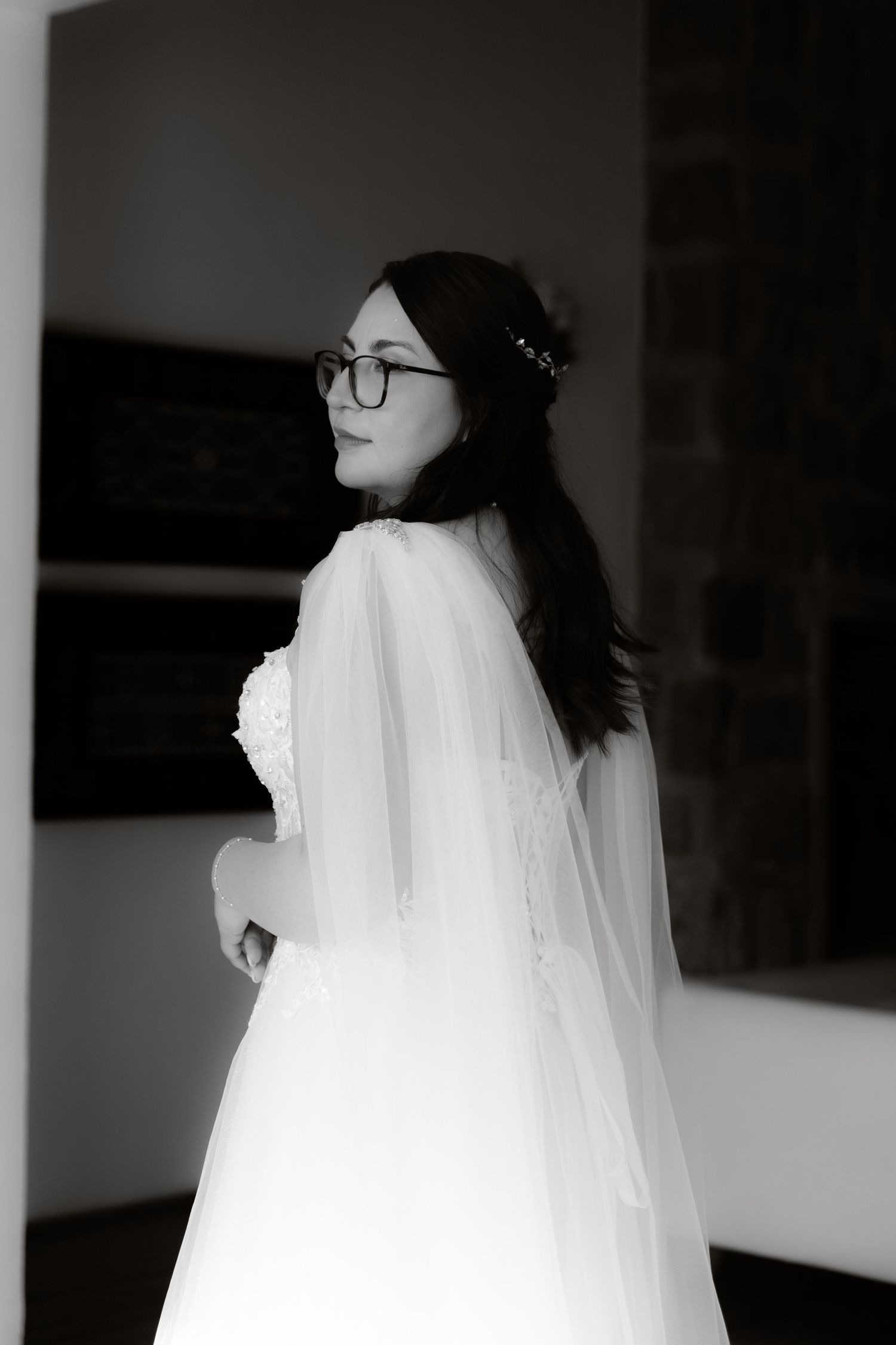 Black and white portrait of bride in lace wedding dress and glasses with flowing cape veil.