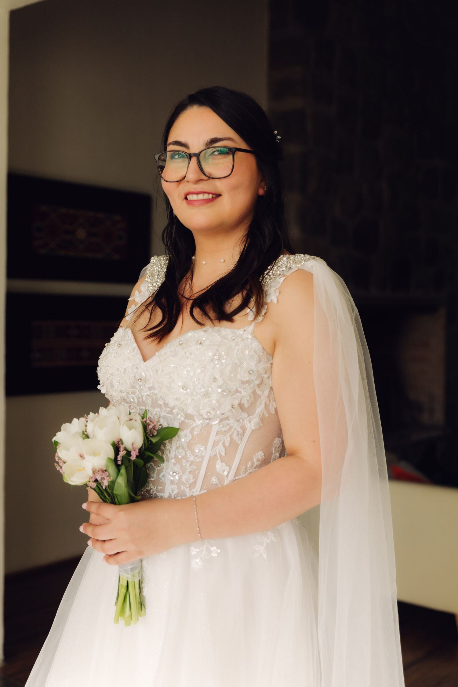 Smiling bride in beaded lace wedding dress holds white bouquet while wearing glasses indoors.