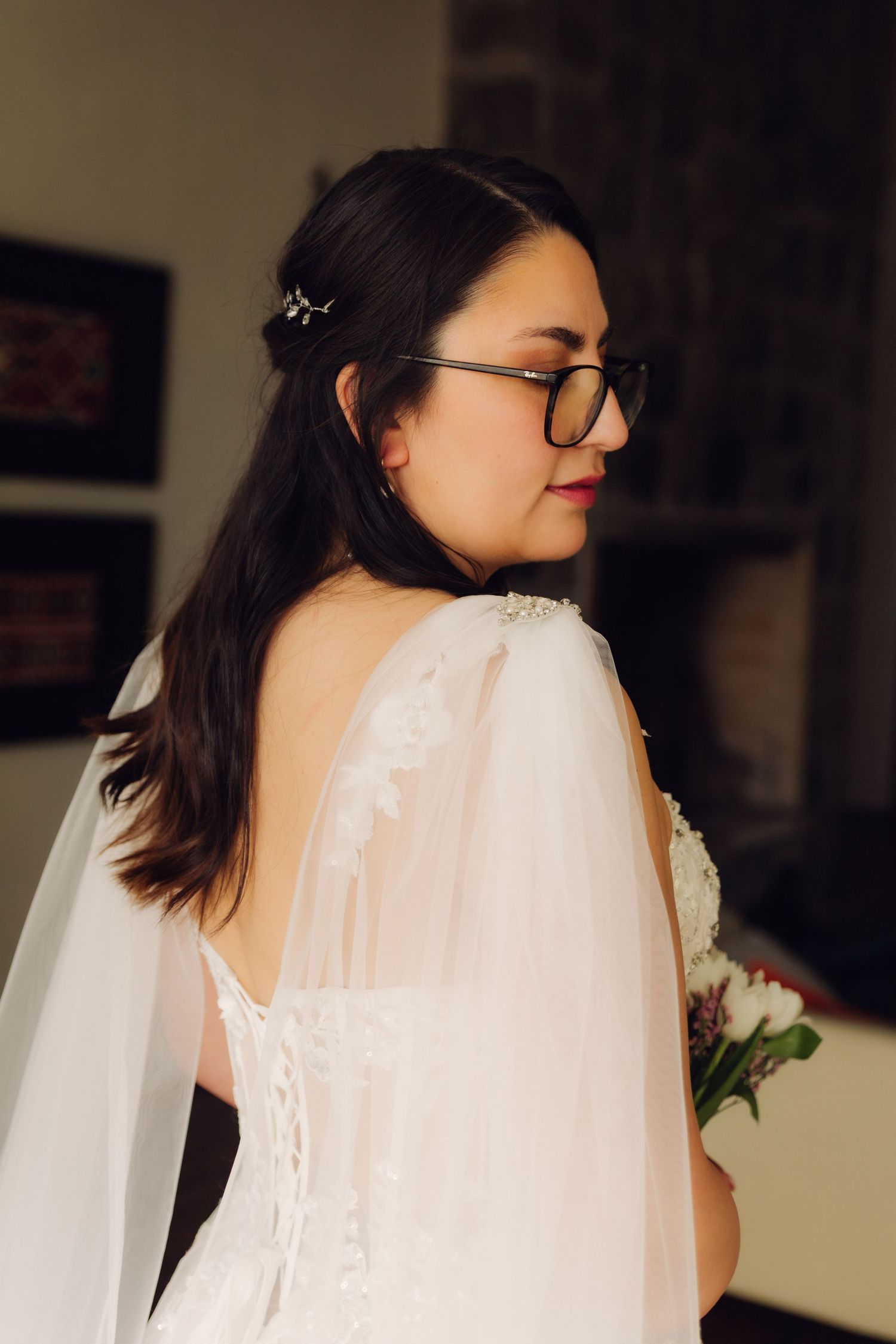 Bride in white wedding gown with cape veil and glasses, looking over shoulder indoors.