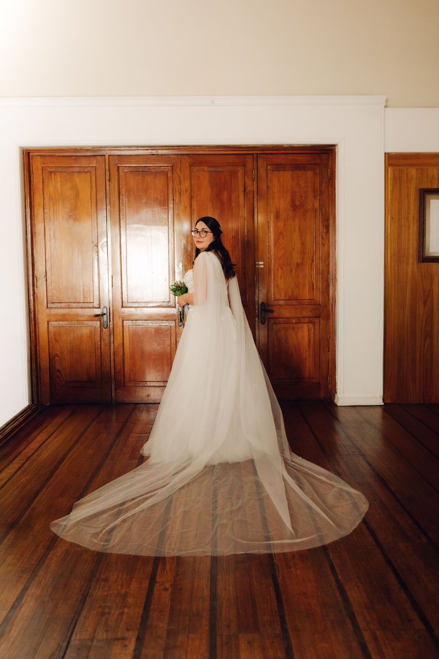 Full-length bridal portrait showing bride in white gown with long cathedral cape veil by wooden doors.