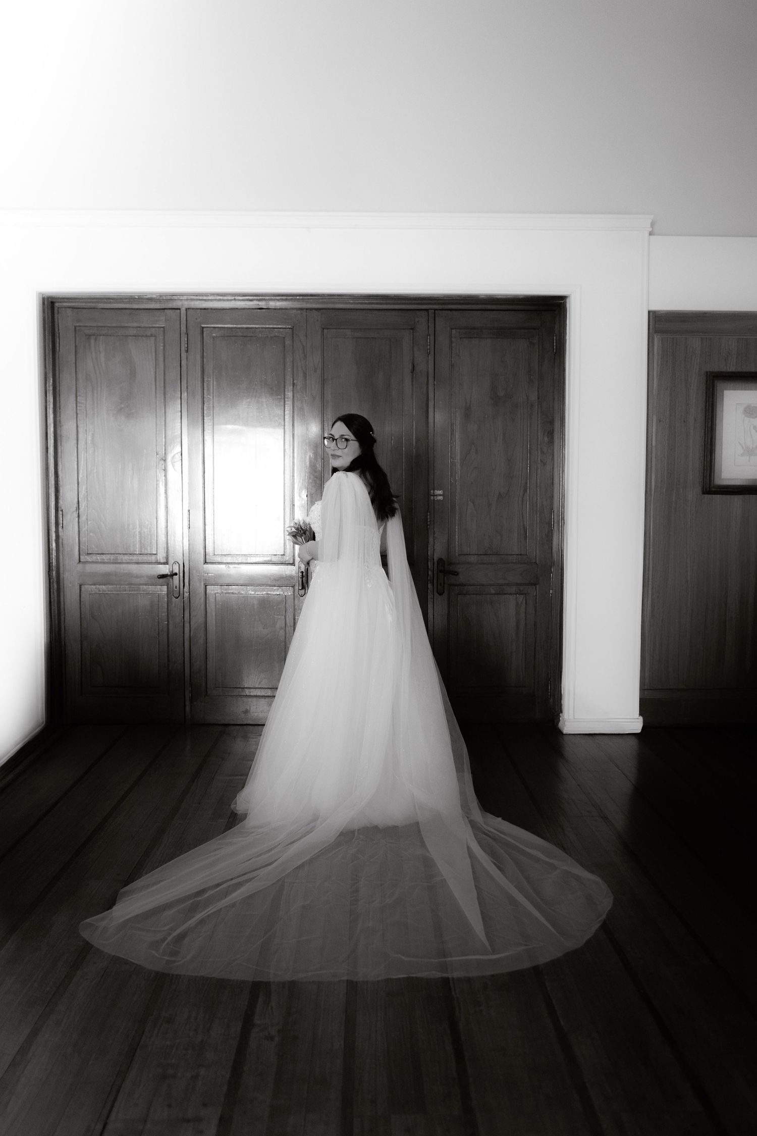 Black and white photo of bride in full-length wedding dress with long train standing by wooden doors.