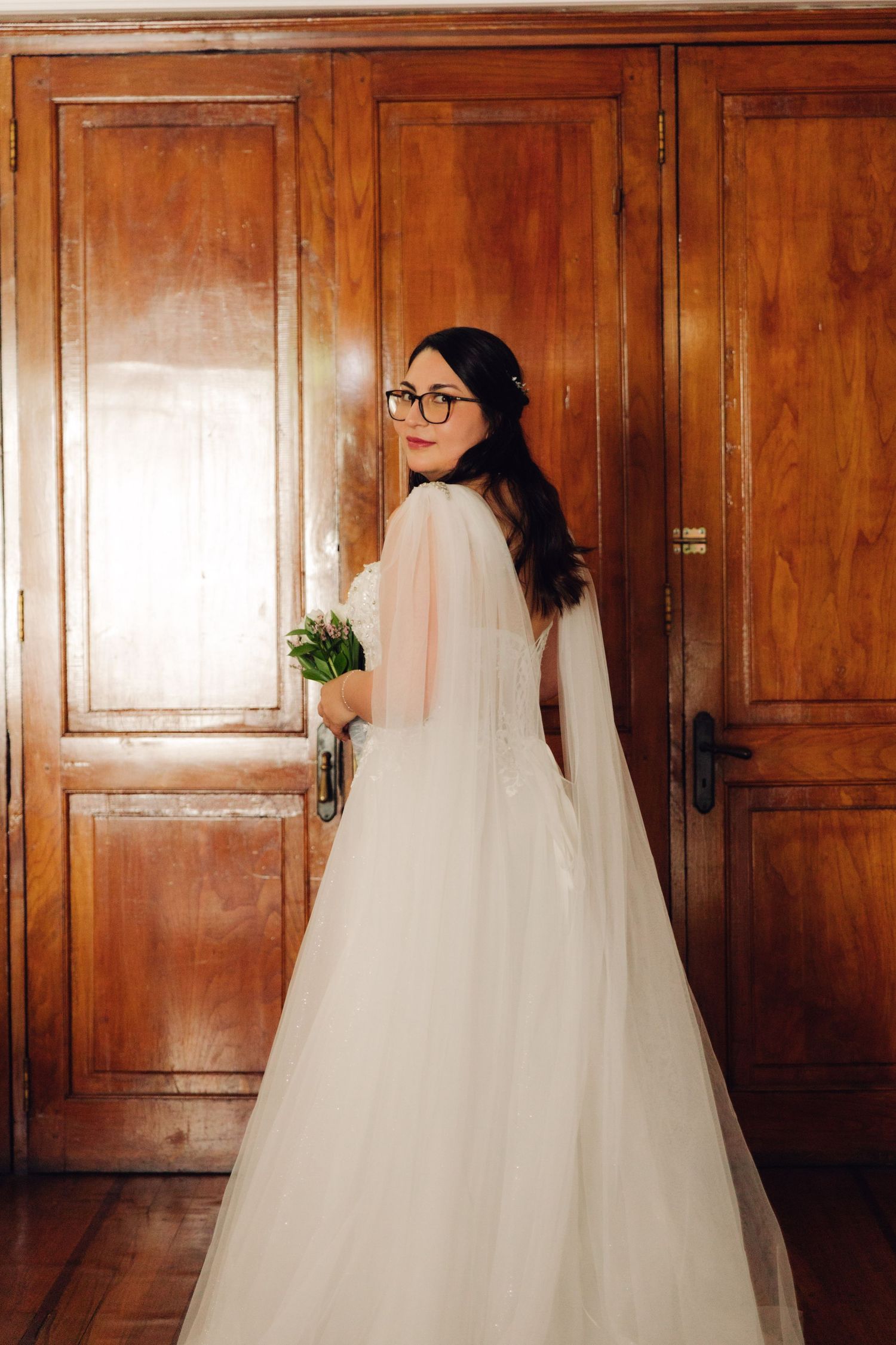 Smiling bride in white wedding dress with cape and glasses holding bouquet by wooden doors.