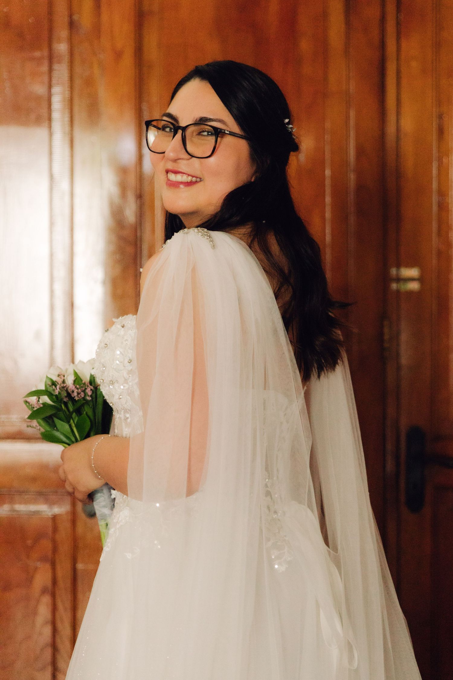 Radiant bride in white wedding dress with sheer cape and glasses smiling over her shoulder.