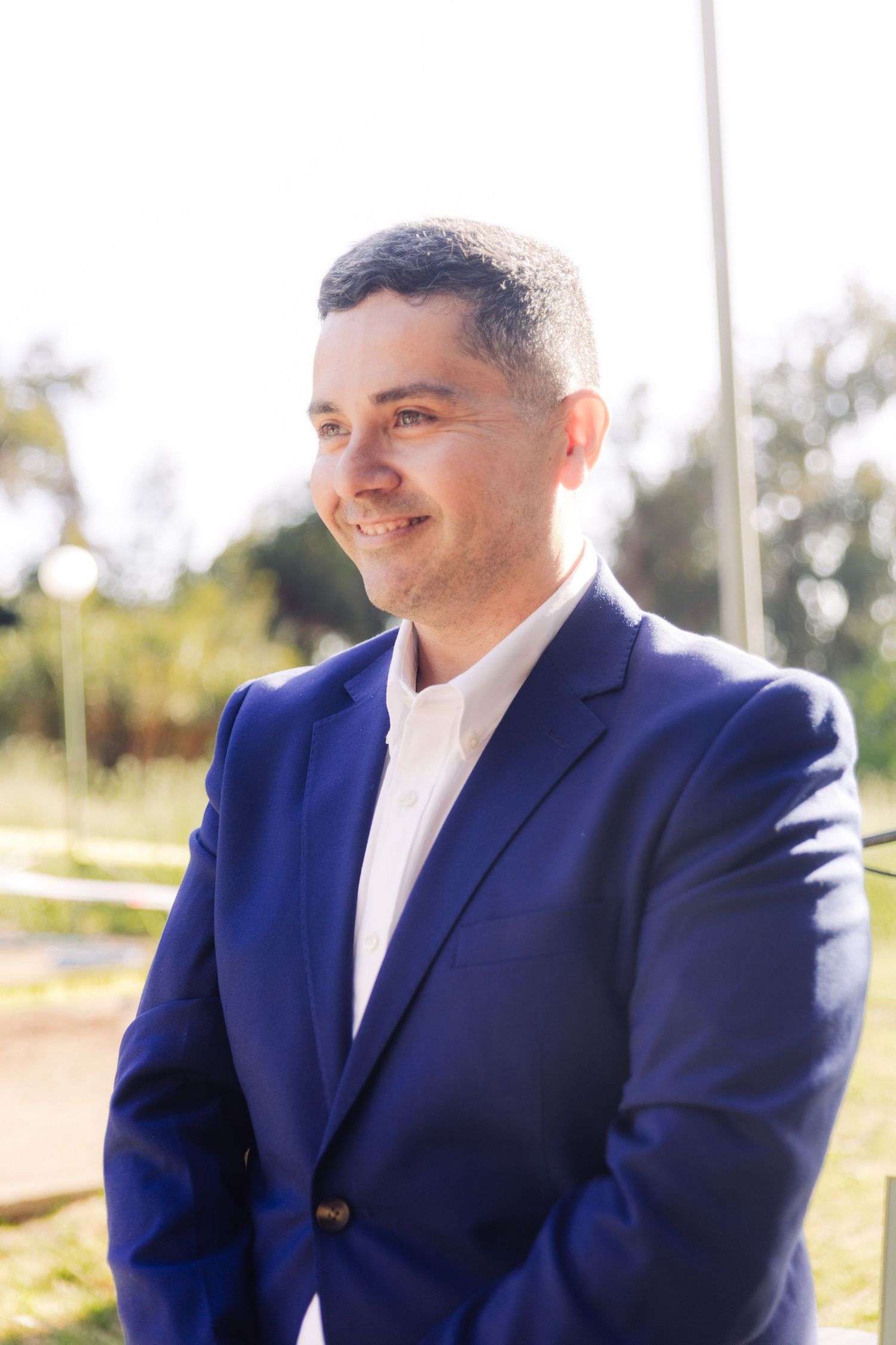 Man in blue blazer and white shirt smiling outdoors with greenery in background.